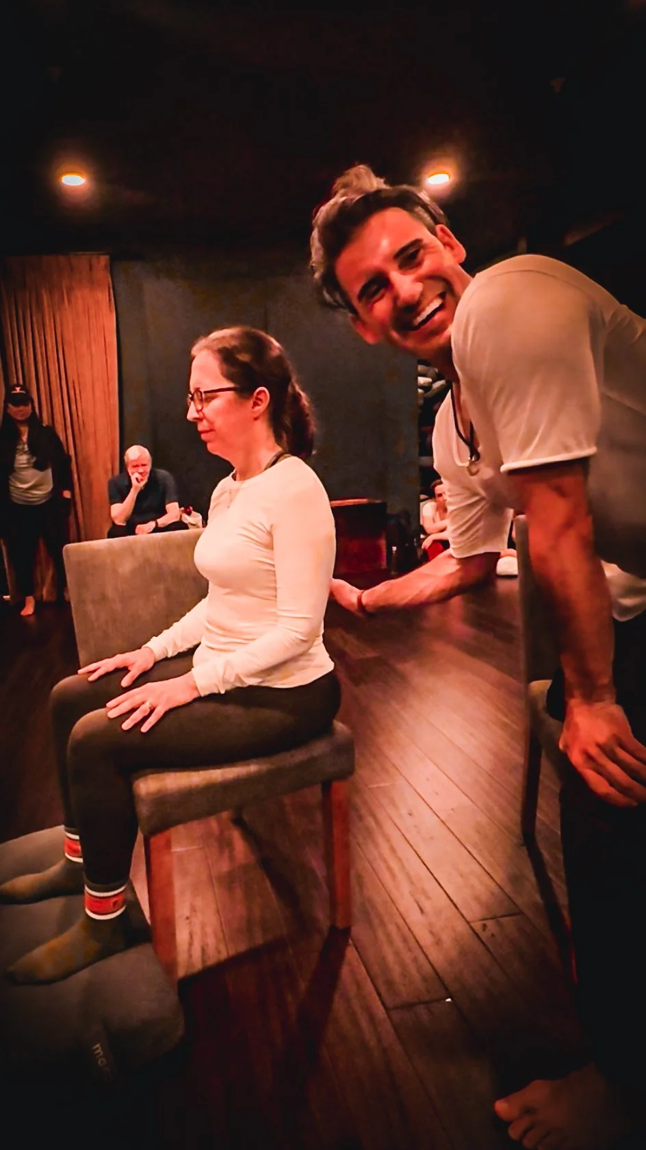 A man with dark hair smiling and leaning on a table next to a seated woman with glasses, in a dimly lit room, with other people in the background.