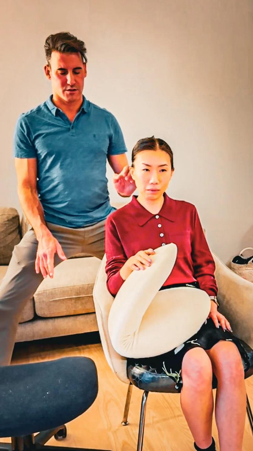 A man with dark hair wearing a blue polo shirt and beige pants demonstrates a physical therapy technique on a seated young woman. The young woman has dark hair in braids, wearing a red blouse and a black skirt, with her leg bent and resting on a chair.