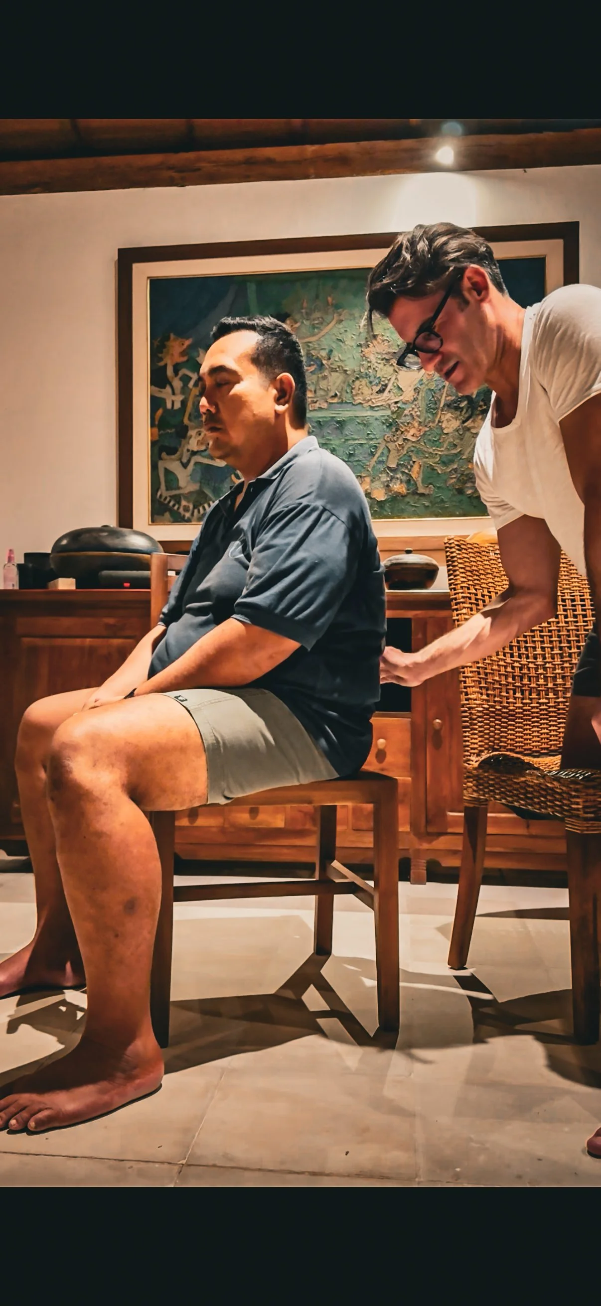 A man sitting on a wooden chair with his eyes closed, receiving a massage from another man standing behind him. The setting appears to be a room with wooden furniture and a decorative painting on the wall.