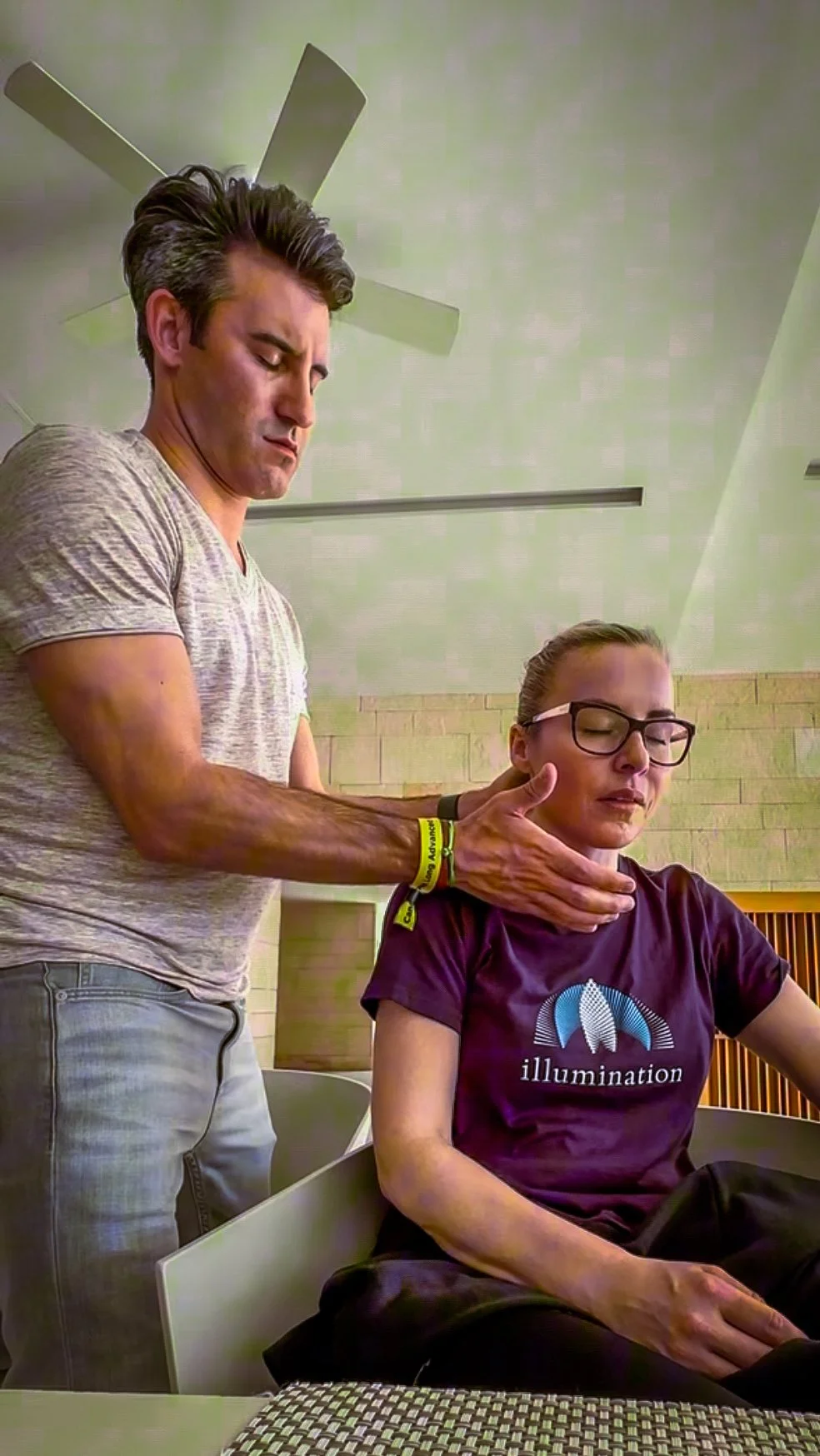 A man performs a neck exercise or stretch on a woman who has her eyes closed and appears relaxed, in a room with a ceiling fan and beige walls.