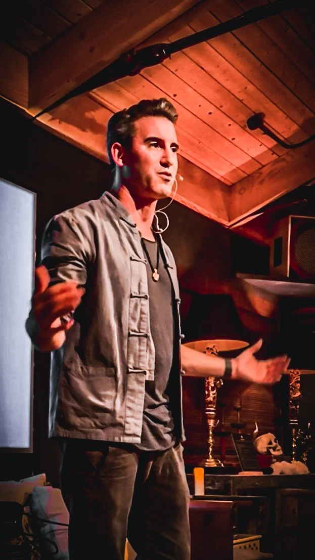 Young man speaking or performing on stage with a microphone, gesturing with his arms, in a cozy, wood-paneled room with artistic items and a skeleton on a table in the background.