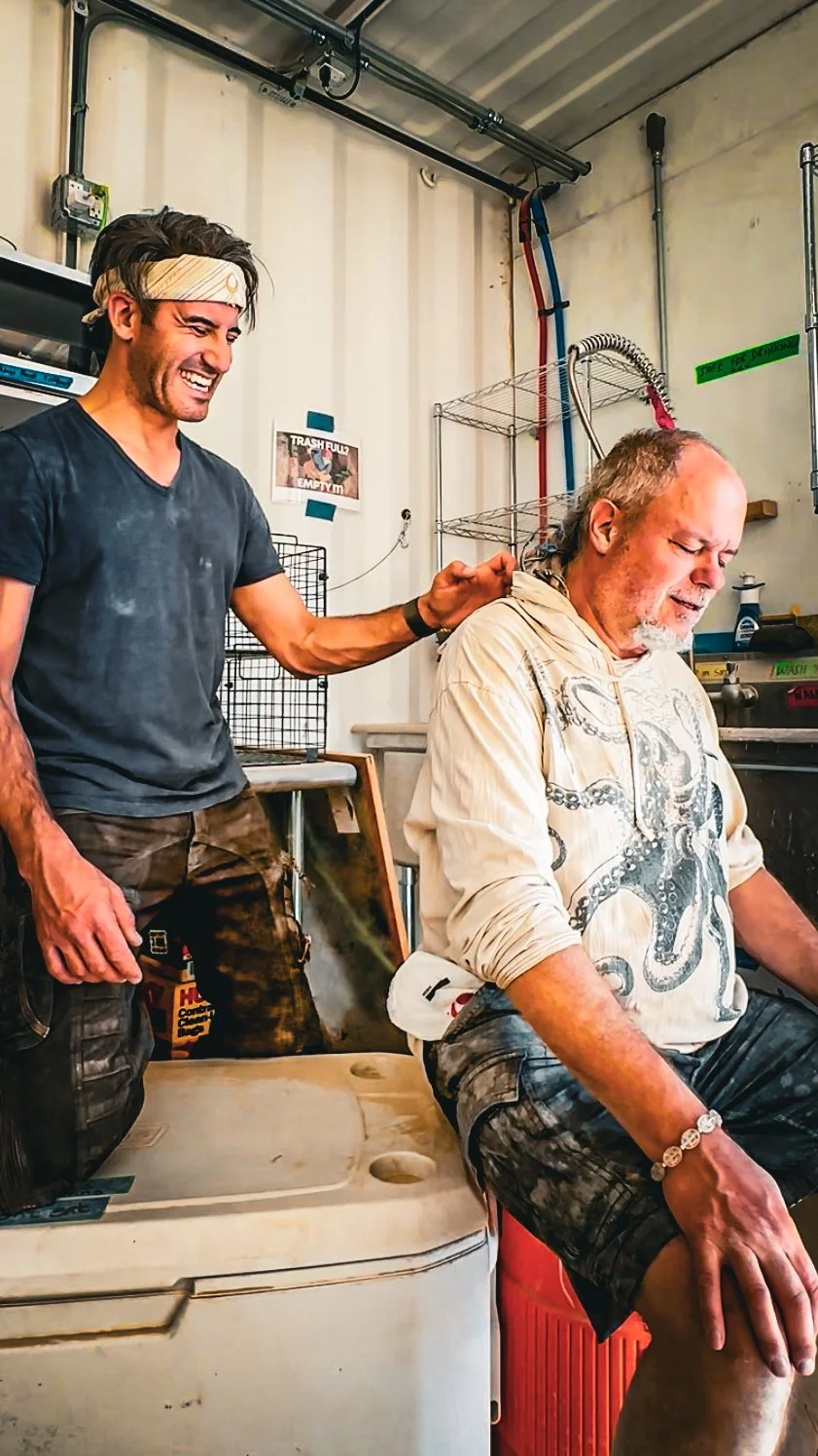 Two men in a garage or workshop, one younger with dark hair and a headband, smiling and touching the other older man who is sitting on a cooler, with a distressed expression. The background includes wire racks, pipes, and signs.
