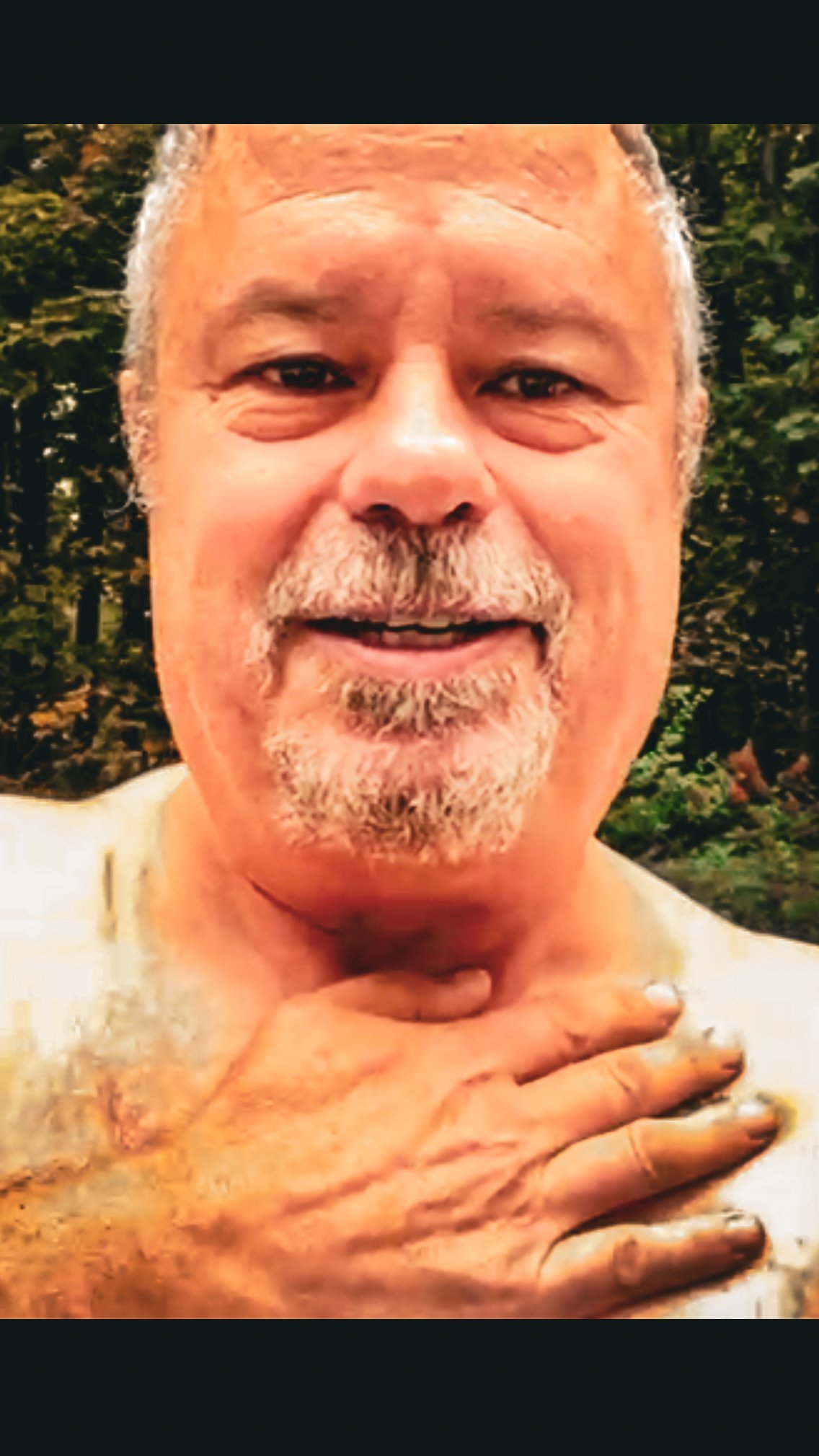 A close-up of an older man with a gray beard and mustache, smiling at the camera, with his hand gently placed on his neck. The background is outdoors with trees.