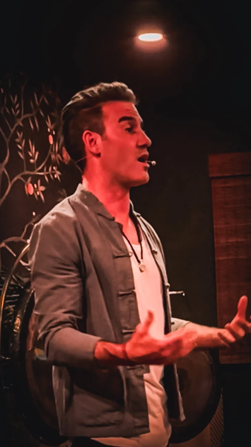 A man speaking or singing with a microphone headset in a dimly lit room with dark walls, decorative plant patterns, and a wooden panel.