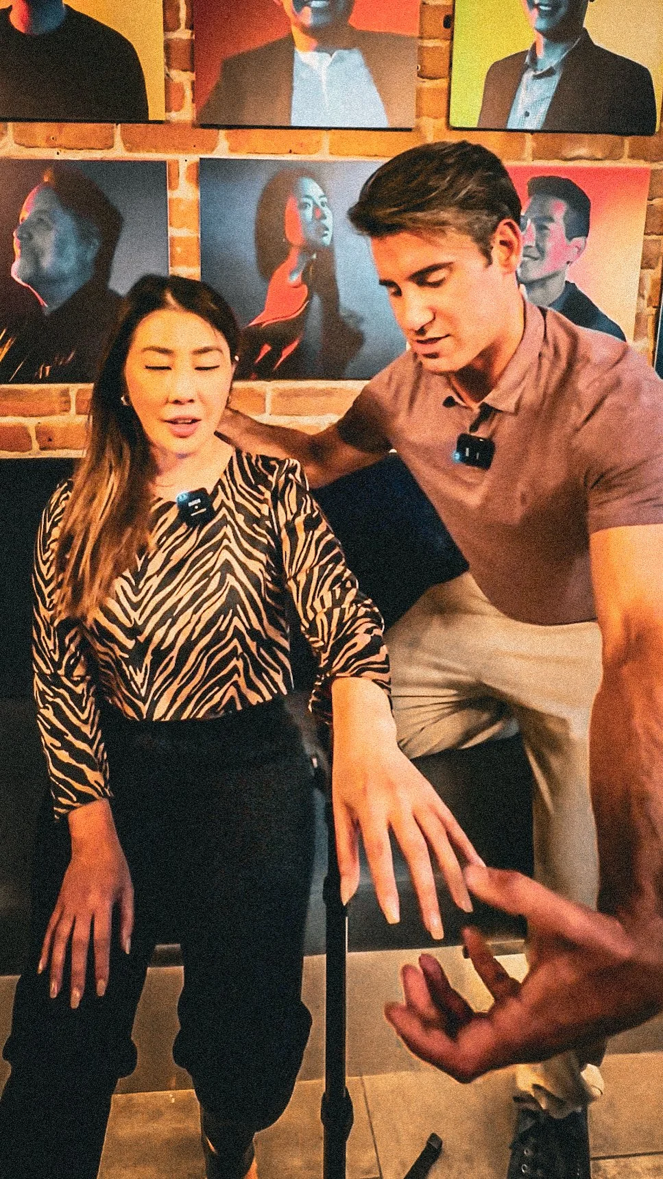 A woman with long hair and a patterned blouse appears to be experiencing a medical issue, with her eyes closed and her hand extended. A man in a brown shirt next to her is reaching out and speaking, with a person in the foreground gesturing with their hand. The background features a brick wall with colorful portrait paintings of various people.