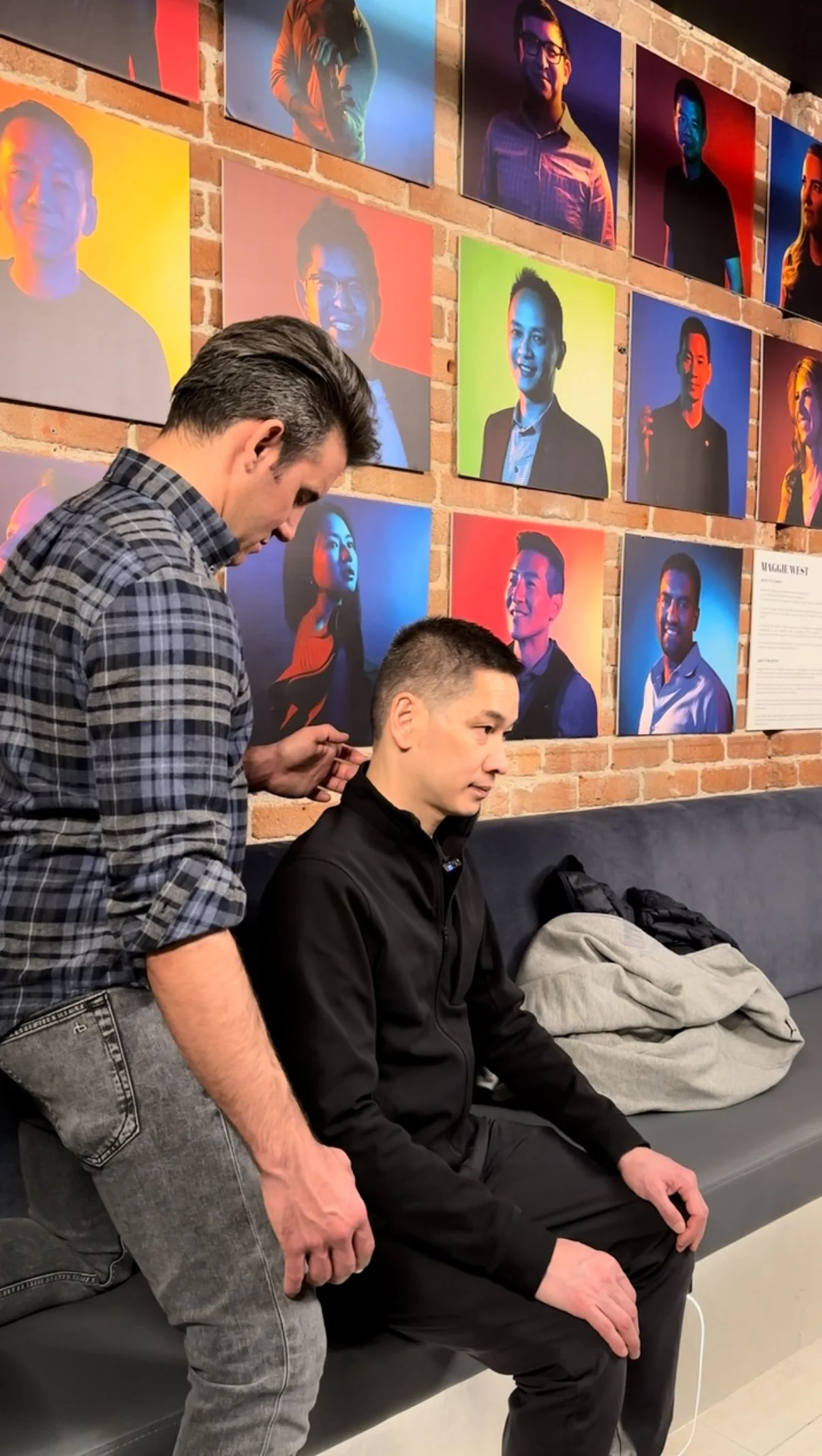 A man with short black hair, wearing a black jacket, sitting on a black bench. Another man with dark hair, wearing a plaid shirt, standing next to him. They are in an art gallery with a brick wall decorated with colorful portrait photographs of diverse people.