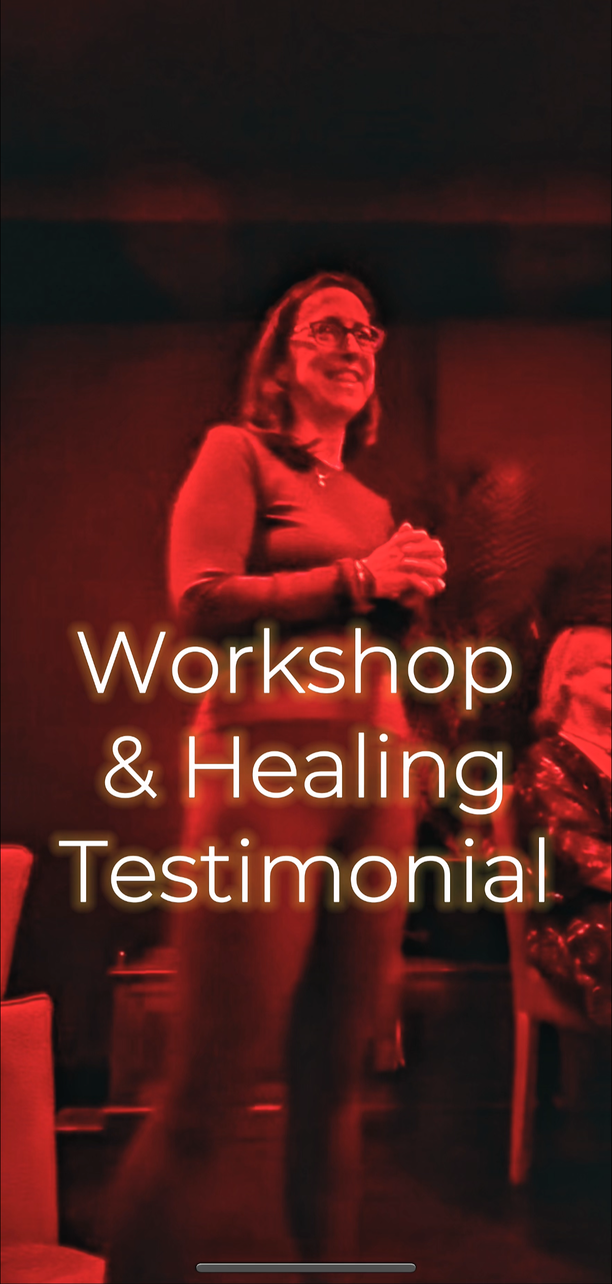 A woman standing, smiling, with glasses, at a workshop and healing testimonial event, with a blurred background.