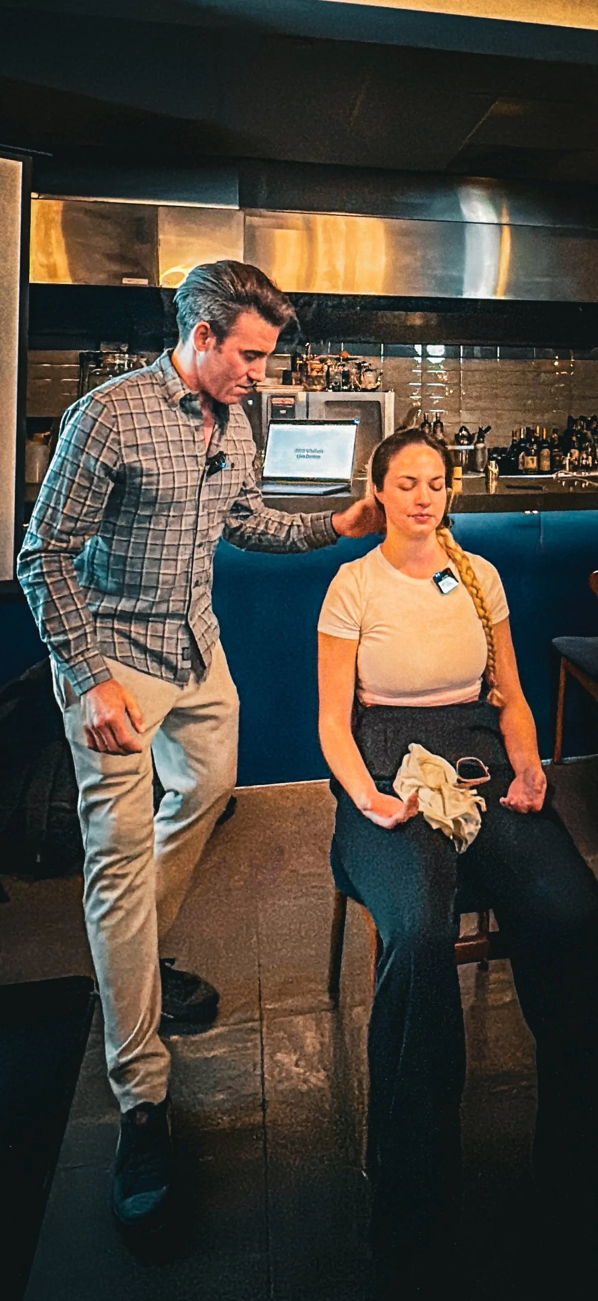 A man is standing beside a seated woman in a bar or restaurant setting, appearing to check her neck and back. The woman has her eyes closed and a towel or cloth on her lap, with a glass of wine in her hand.