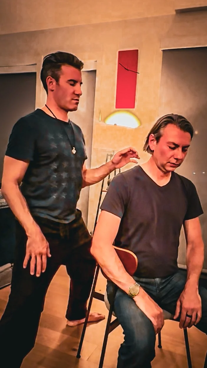 Two men, one sitting and one standing, in a dimly lit room with wooden floors and beige walls. The standing man appears to be giving a massage or performing a healing touch on the seated man's shoulder.