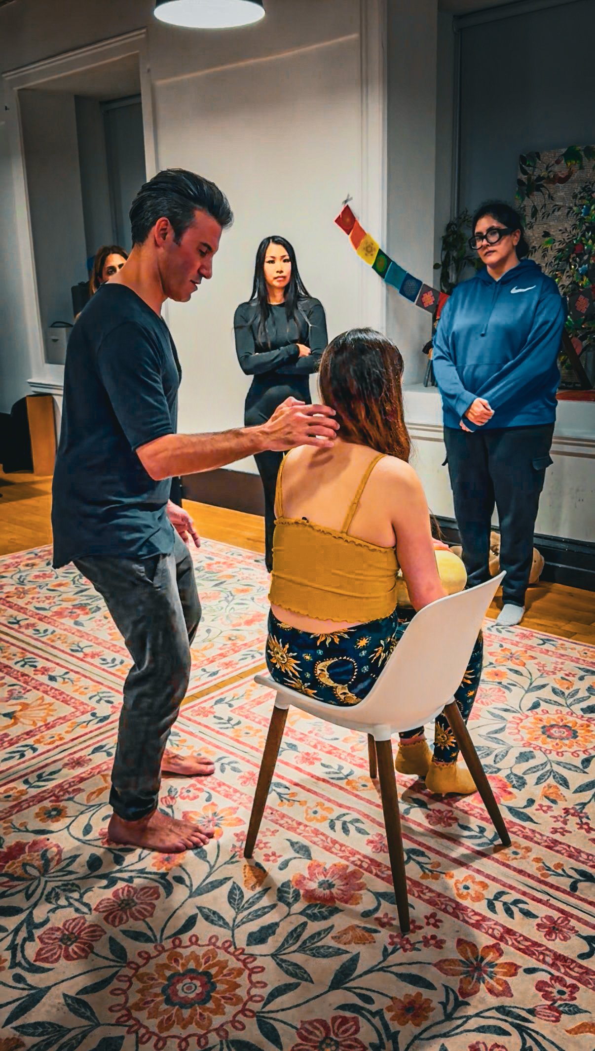 A woman is seated on a white chair on a colorful patterned rug, with three women standing behind her and a man standing beside her touching her head. One of the women standing is wearing a blue hoodie, another in black, and the woman seated is wearing a yellow top and floral pants. The scene appears to be a spiritual or healing session taking place indoors.