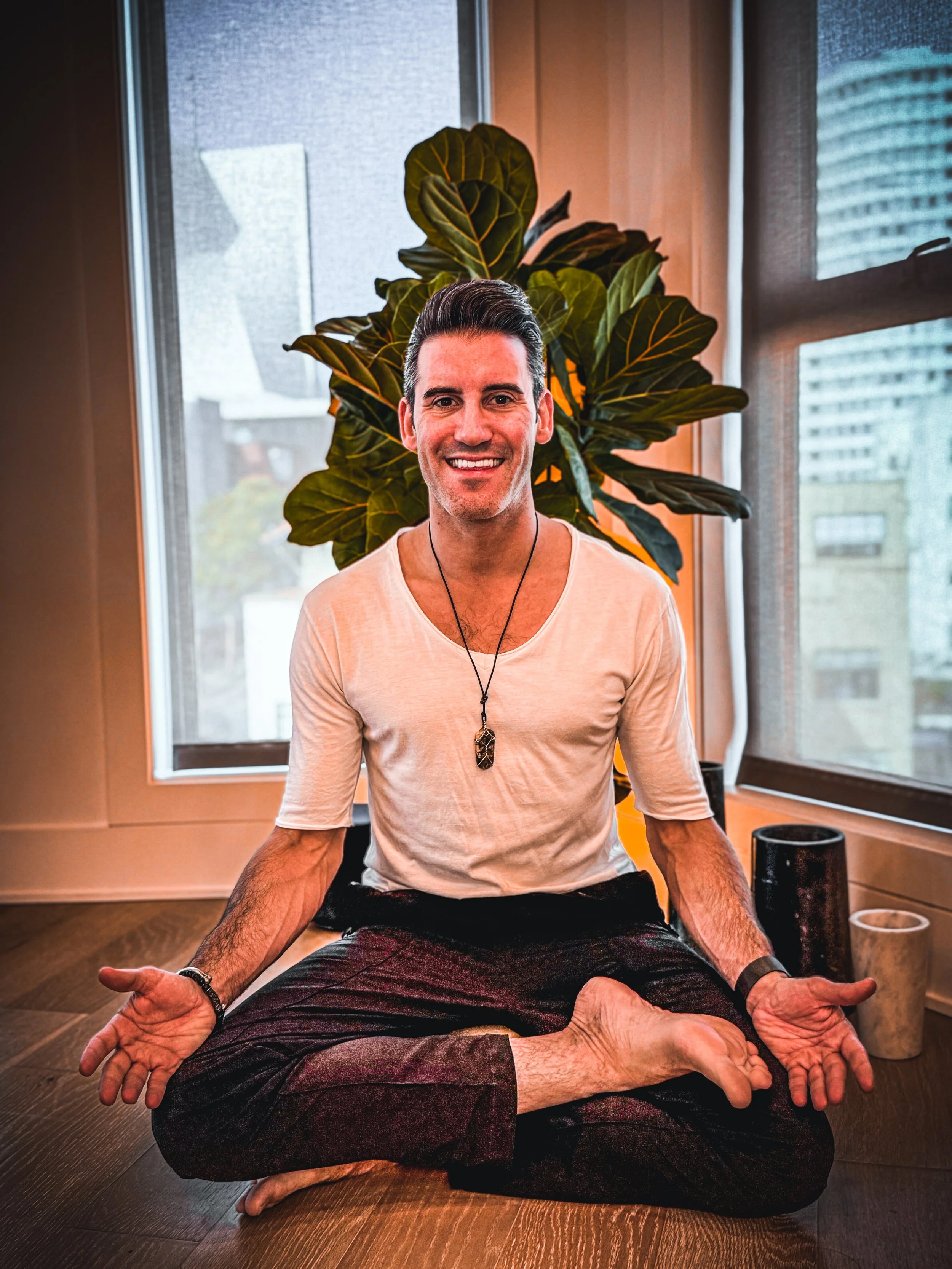 A man practicing yoga or meditation in a seated cross-legged position indoors with large windows and a plant behind him.