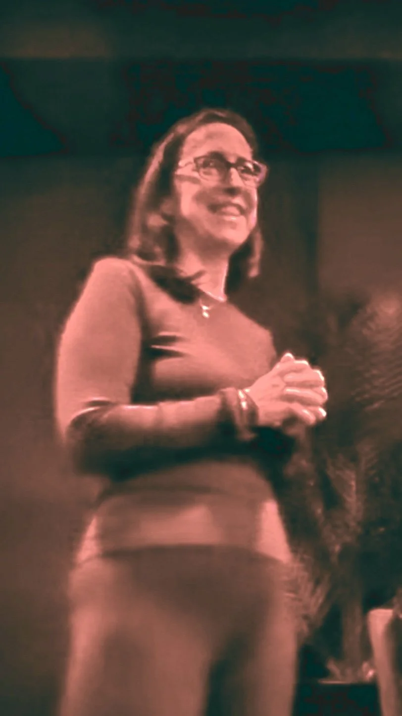 A woman with glasses, shoulder-length hair, wearing a long-sleeved top and pants, standing with hands clasped, smiling in a dimly lit indoor setting.