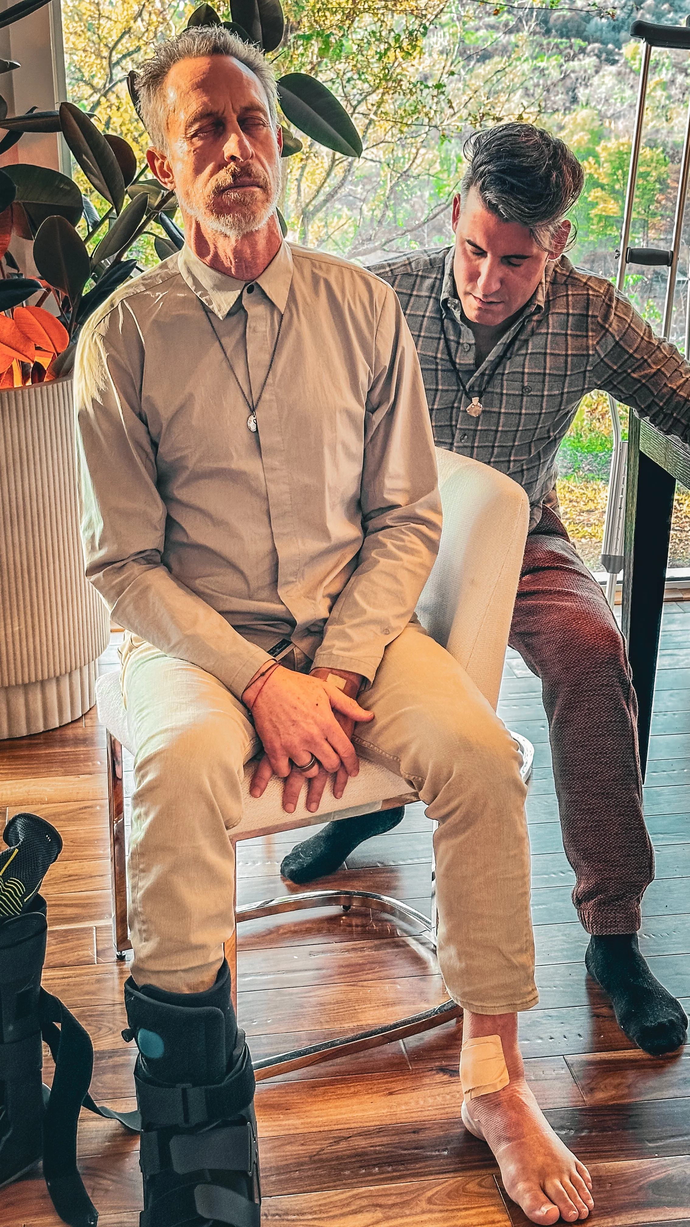 An older man sitting on a chair with his eyes closed, wearing a beige shirt and light-colored pants. He has a bandage on his left ankle. A younger man wearing a plaid shirt is standing behind him, leaning over and looking down at the seated man. The setting appears to be a bright room with large windows and plants in the background.