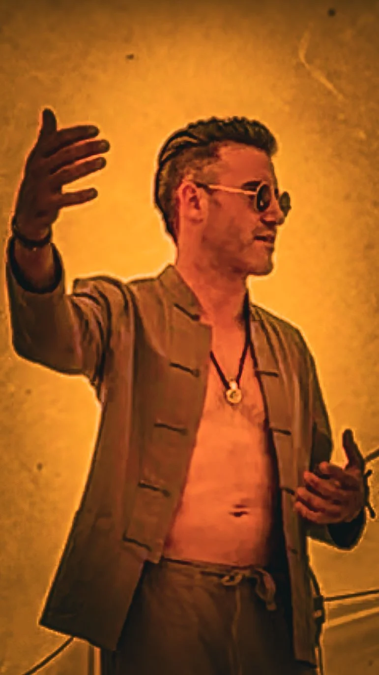 A man with styled hair, wearing sunglasses, a necklace, a tan jacket, and matching pants, is gesturing with one hand, and holding a phone in the other, against a warm yellow background.
