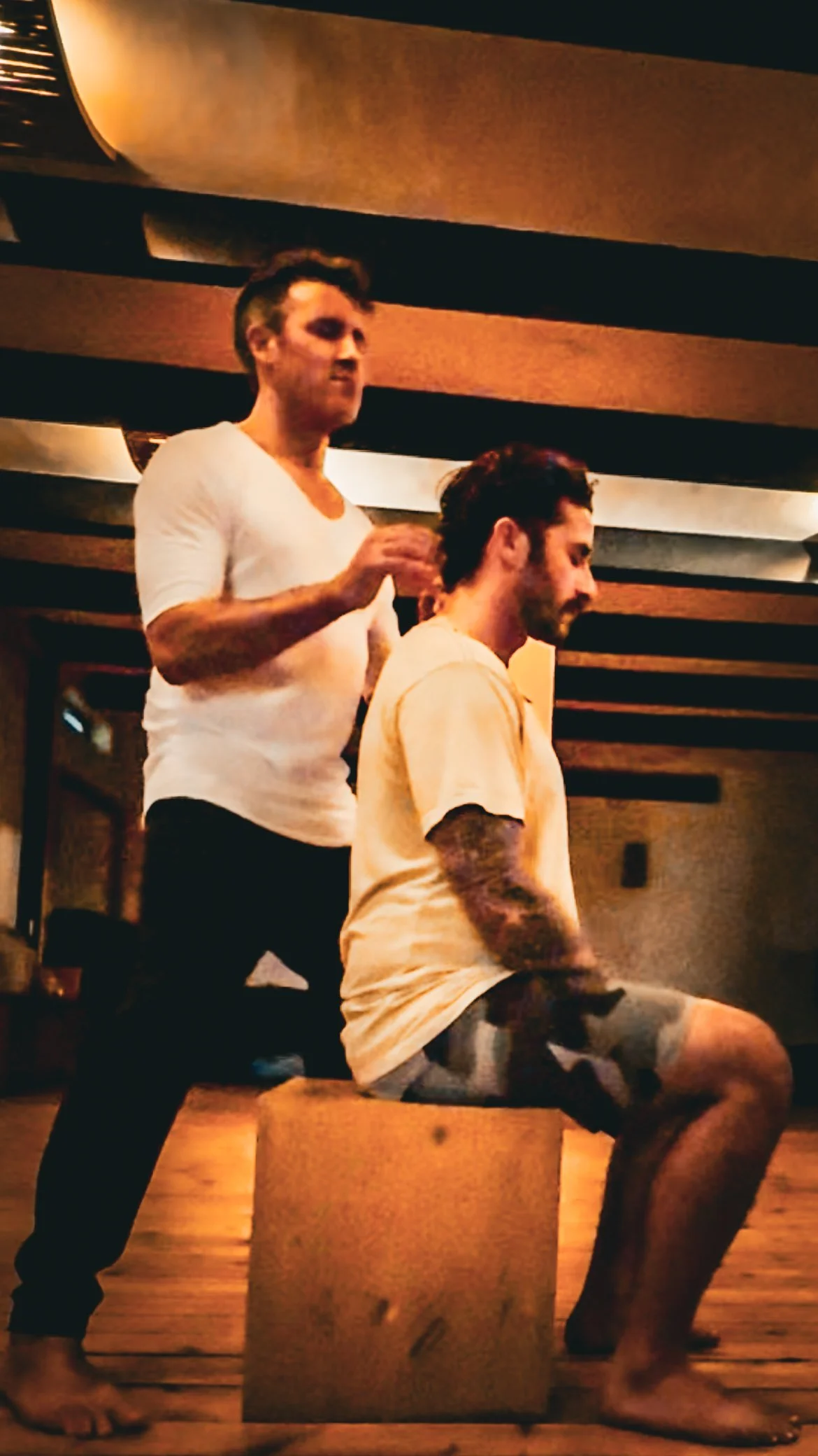 Two men, one sitting on a wooden box and the other standing behind, in a dimly lit room with wooden ceiling panels, possibly in a recording studio or bar.