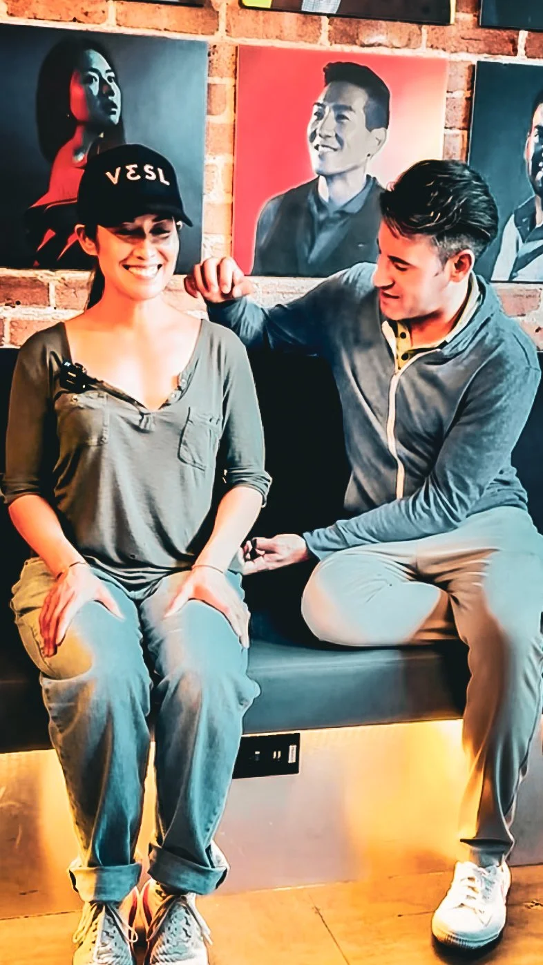 A young woman and a young man sitting on a bench in front of colorful portraits on a brick wall, with the young woman smiling and the young man looking at her.