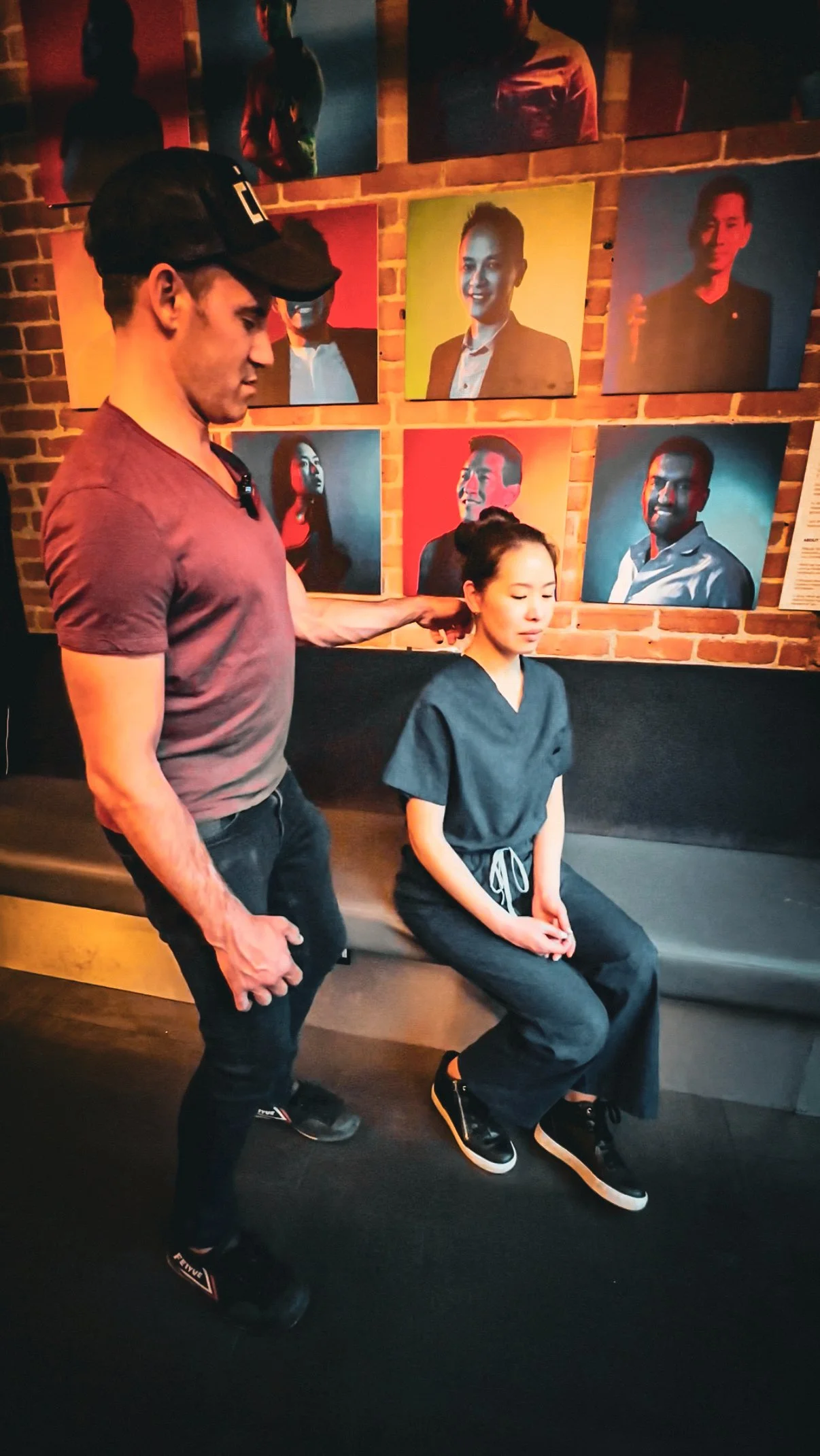 A person in a gray t-shirt and black pants is sitting on a bench with their eyes closed while a male therapist with a black cap and gray t-shirt is standing behind them, gently pulling their head back. The background features a brick wall with colorful portraits of diverse individuals.