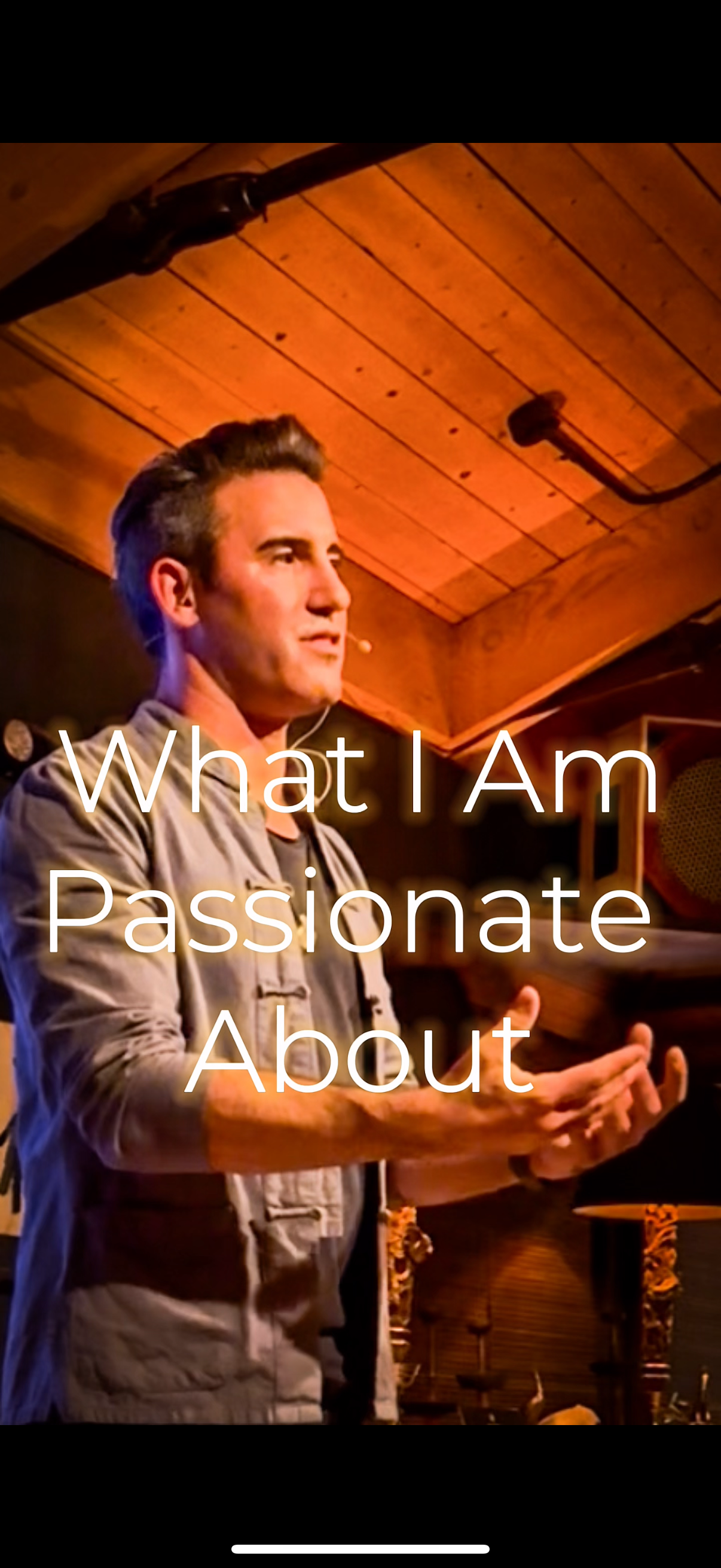 A man with short brown hair, wearing a beige jacket, speaking or presenting in a cozy wooden room with warm lighting.