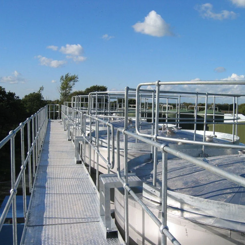 Metal walkway and guardrails surrounding industrial tanks on a rooftop, with a clear blue sky and scattered clouds overhead.