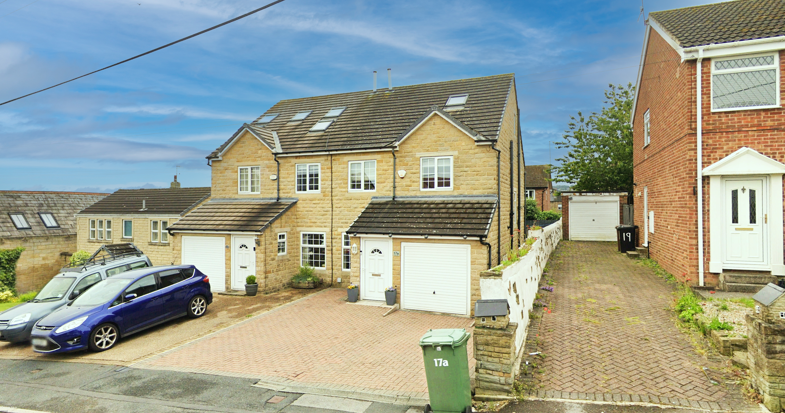 A modern two-story house with a yellow brick exterior, white window frames, and a tiled roof; cars parked in the driveway and a small garden with potted plants. Neighbors' houses connected by a white fence and a paved pathway are visible, with a blue sky overhead.