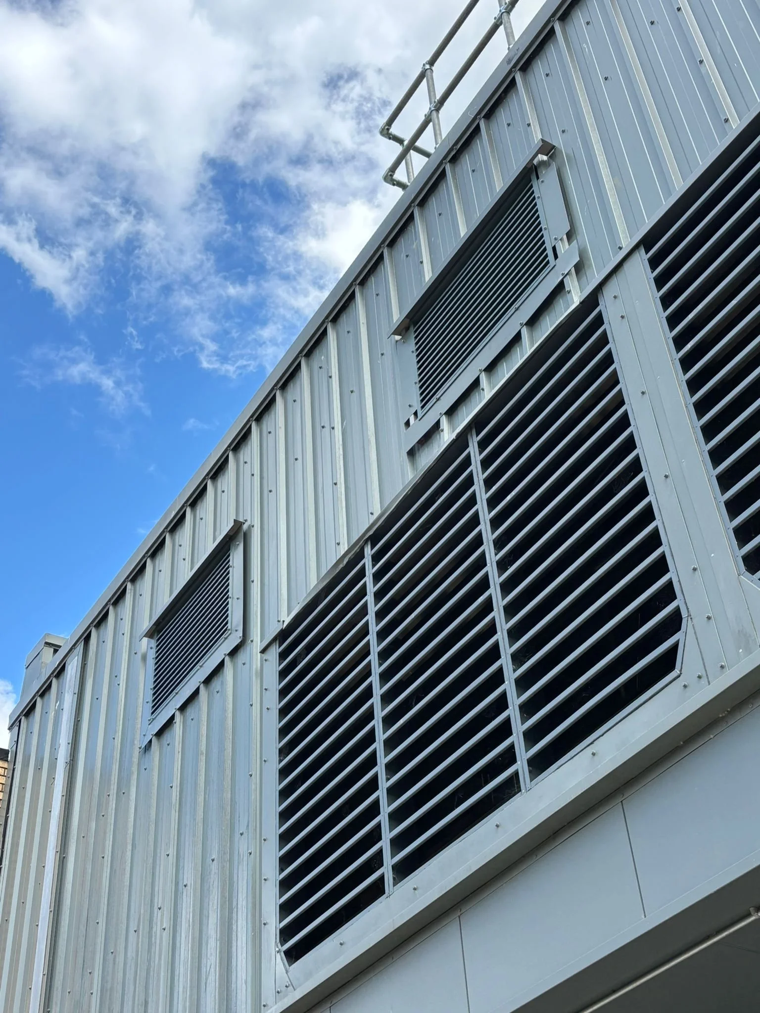 Close-up of a building's exterior with metal siding and large ventilation grilles, against a partly cloudy sky.