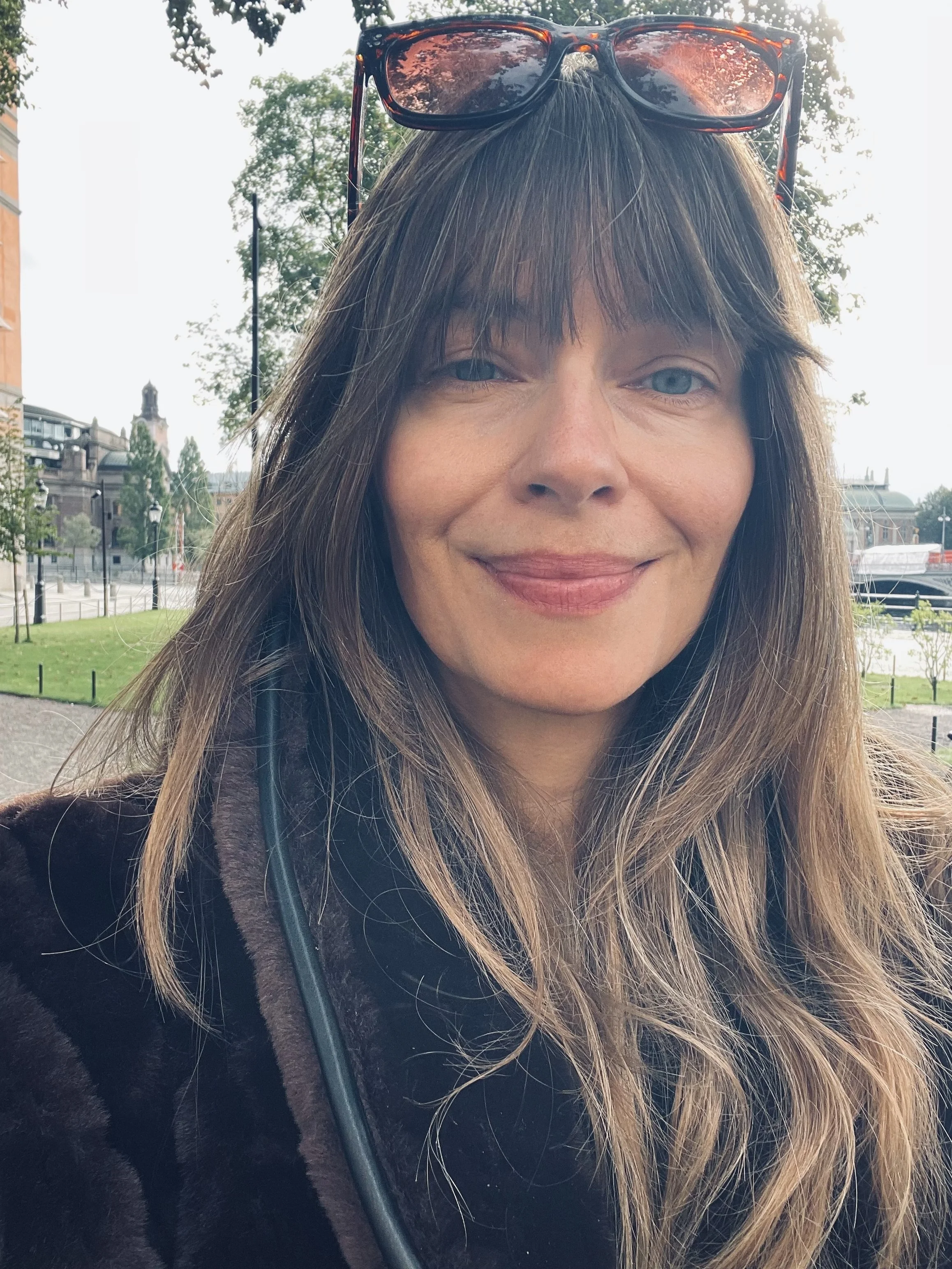 A woman with long brown hair, wearing sunglasses on her head, smiling outdoors near a park and water with buildings and trees in the background.