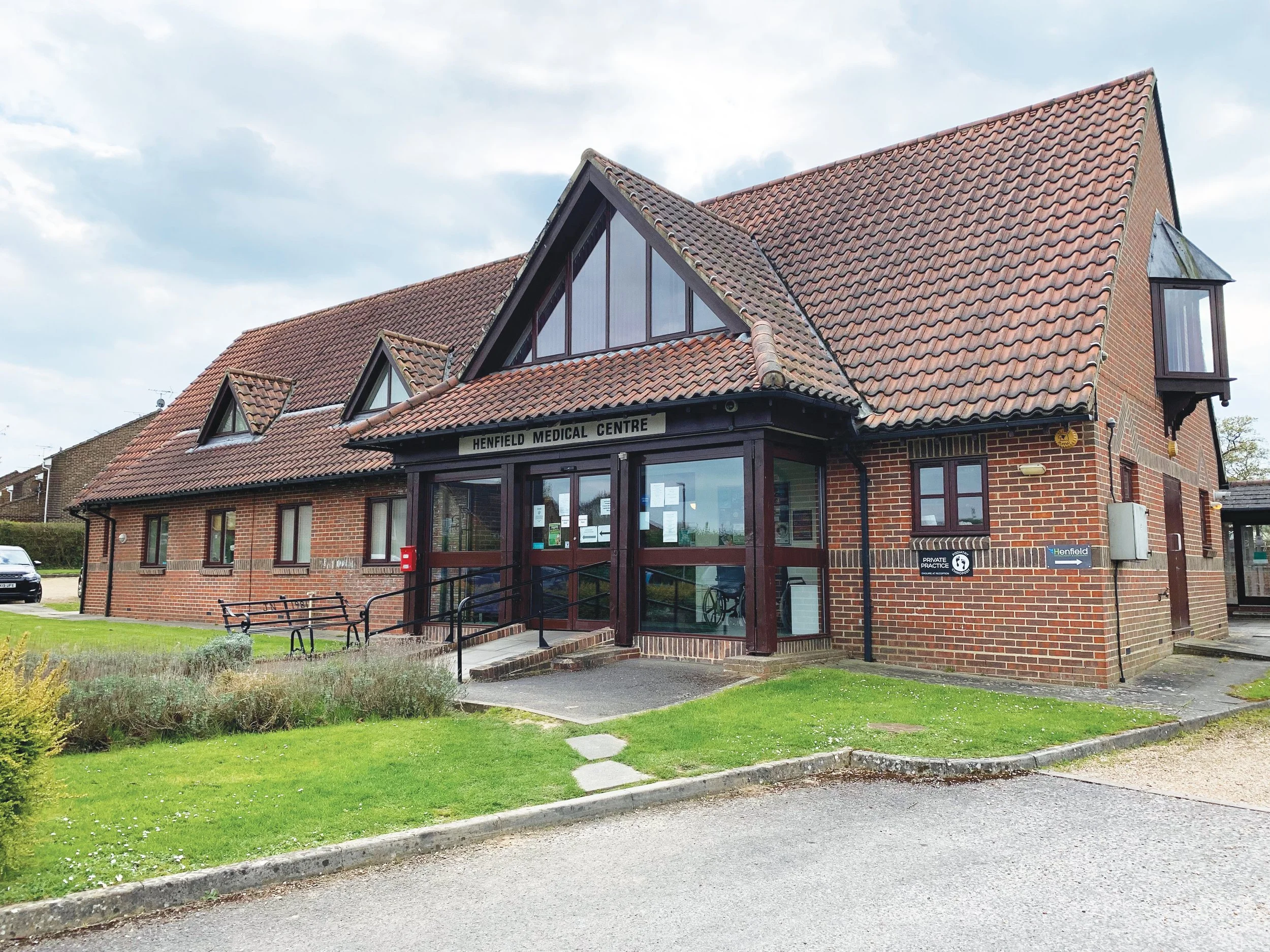 A brick medical centre building with a sign that reads 'Henfield Medical Centre,' entrance with stairs and wheelchair access, and a lawn in front.