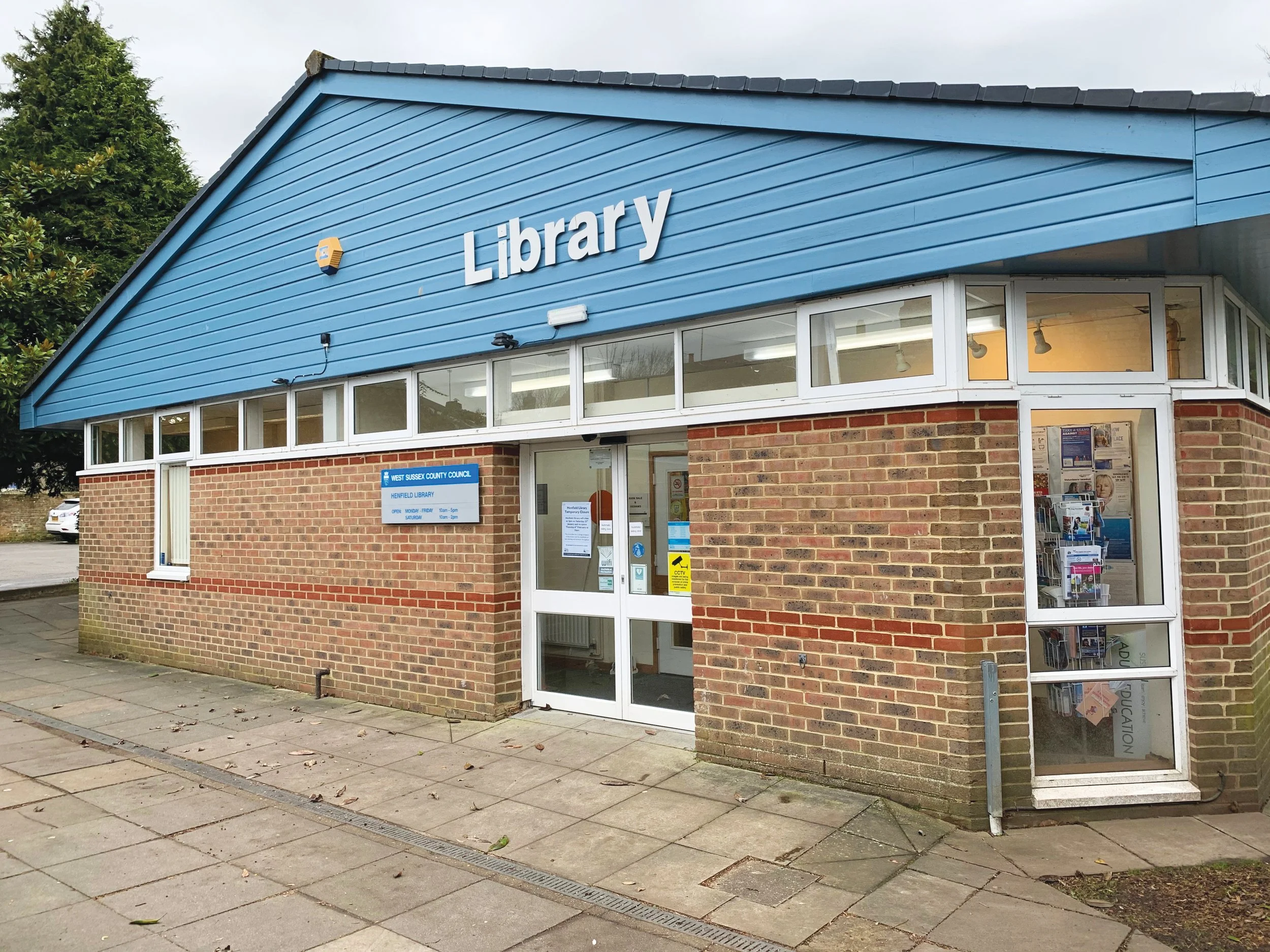 Exterior view of Henfield library building with a blue gable roof and brick walls, front entrance with glass door and window display.
