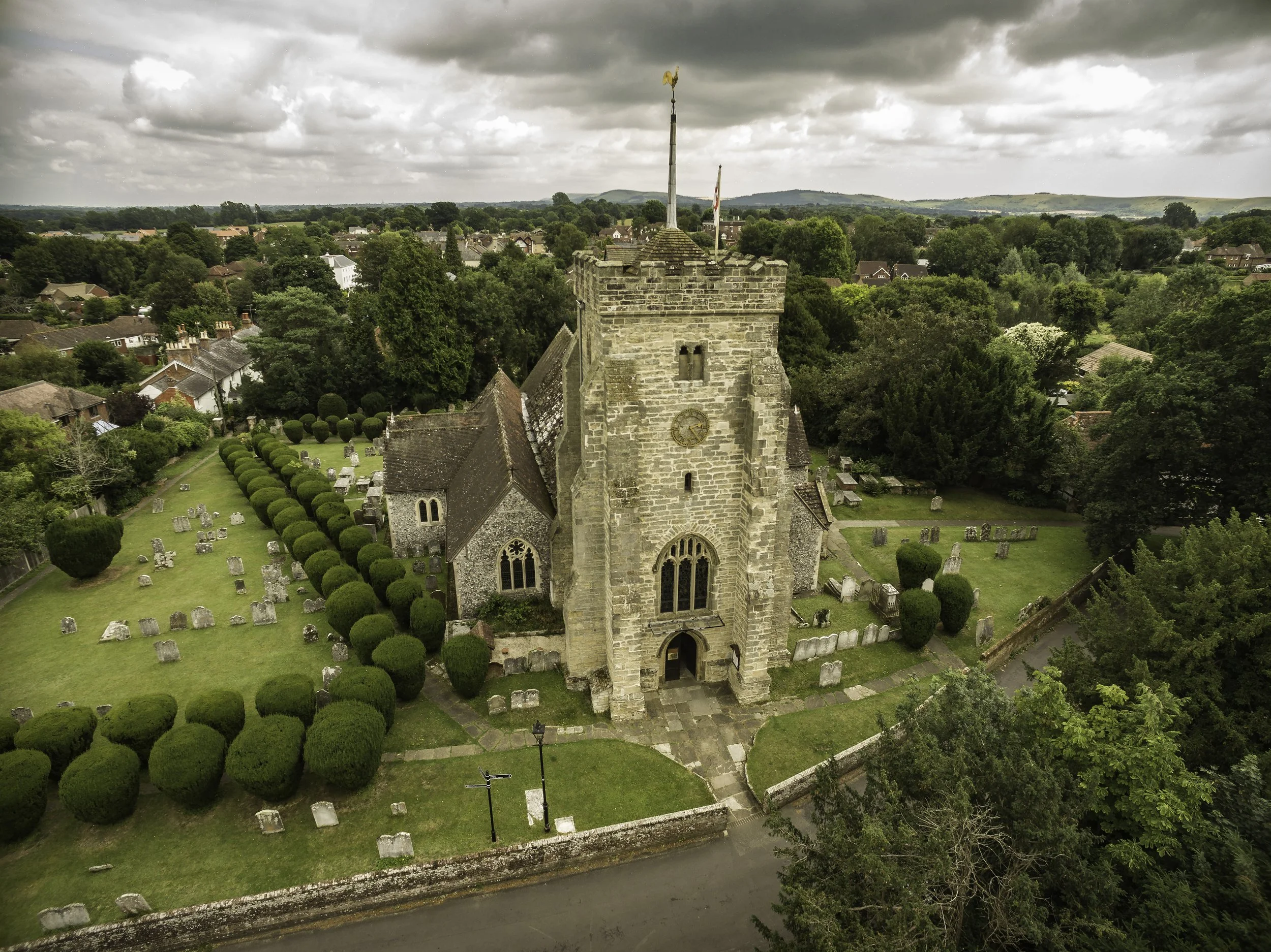 Aerial view of St Peter's Church, Henfield with a clock tower, surrounded by a green churchyard with gravestones and well-maintained hedges, with houses and trees under a cloudy sky.