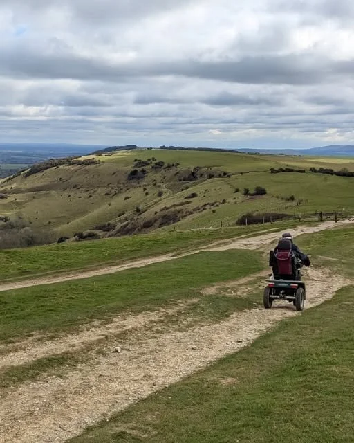 Person riding an electric scooter on a dirt path over the South Downs National Park on a cloudy day.
