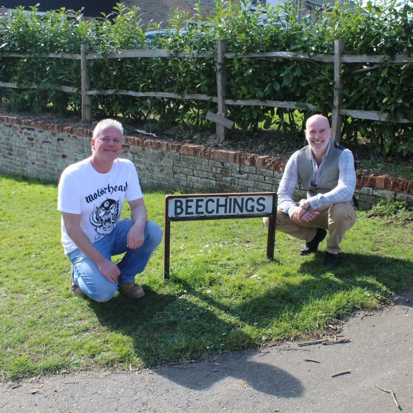 Two of the Tracing the Rails team kneeling on grass near a sign that says 'BEECHINGS' in front of a brick wall and green shrubs.