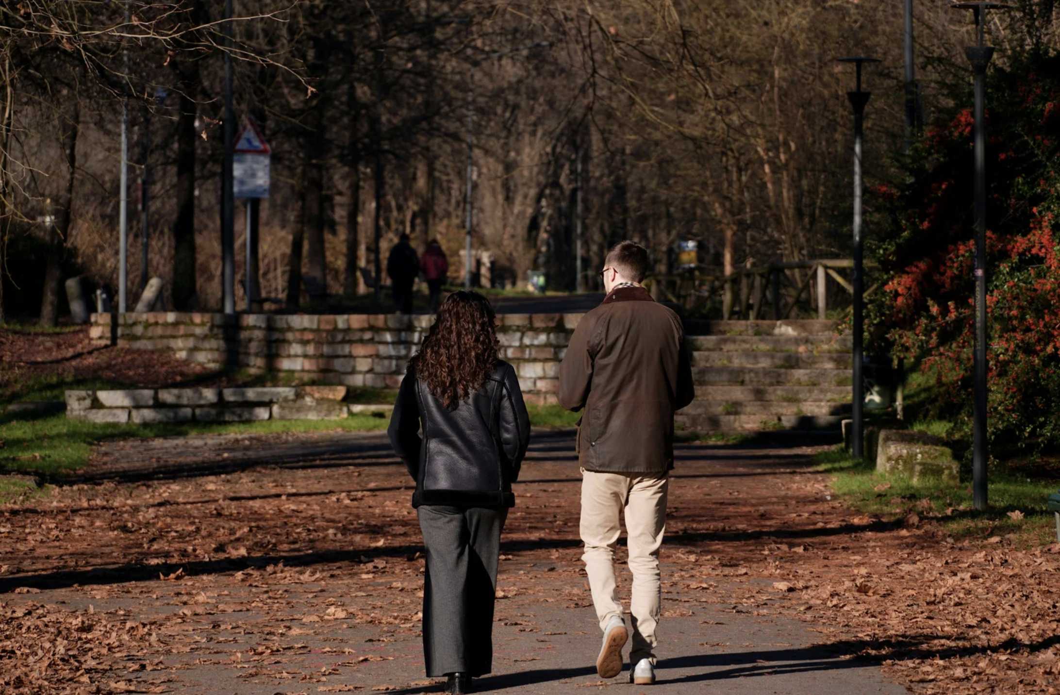 Deux personnes marchant sur un chemin bordé de feuilles mortes, dans un parc avec des arbres et des lampadaires, par une journée en automne.