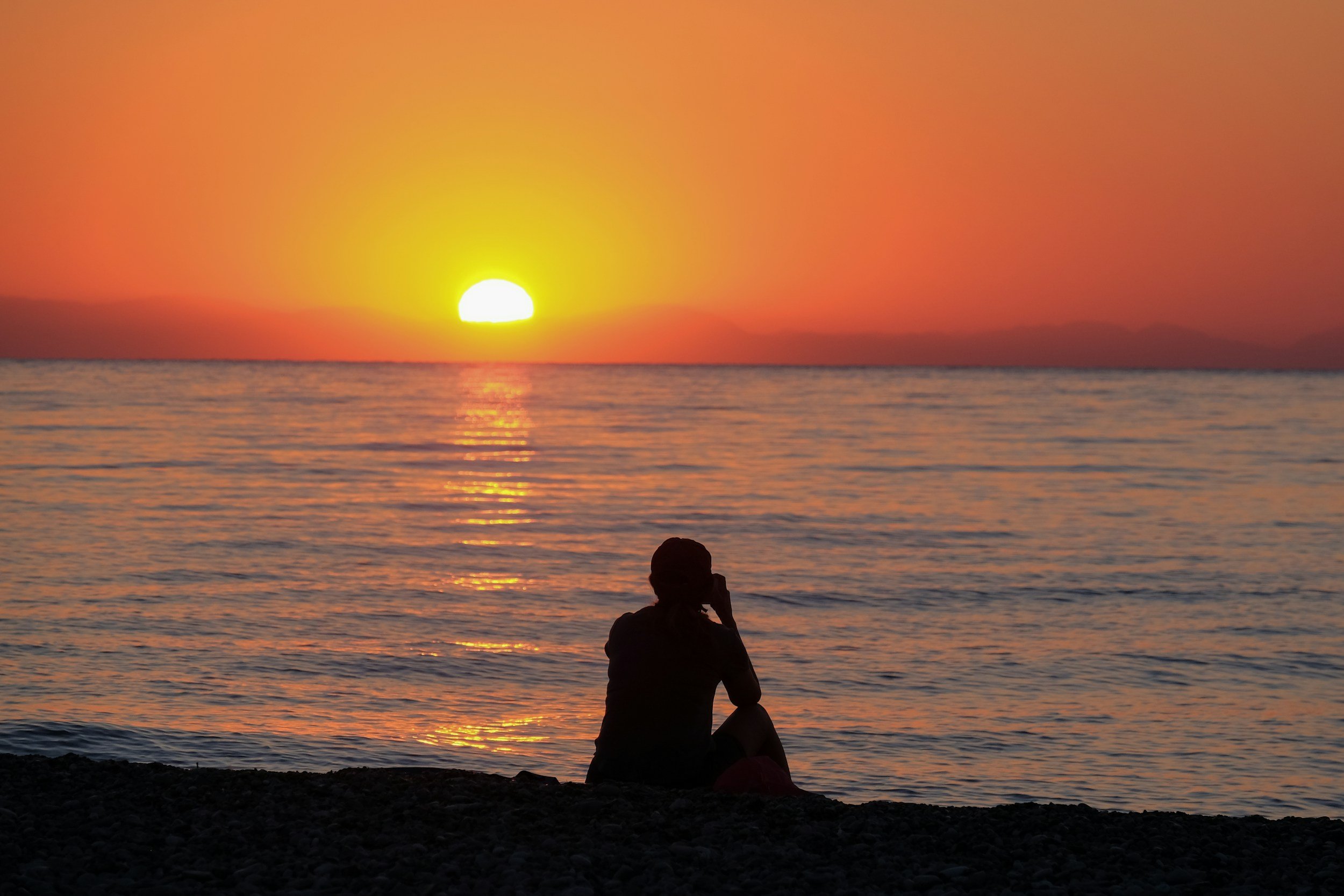 The Open Door - therapeutic practice. Counselling. Therapy for adults. Silhouette of a person sitting on a rocky beach, looking at a sunset over the ocean, with the sun near the horizon and a colourful sky.