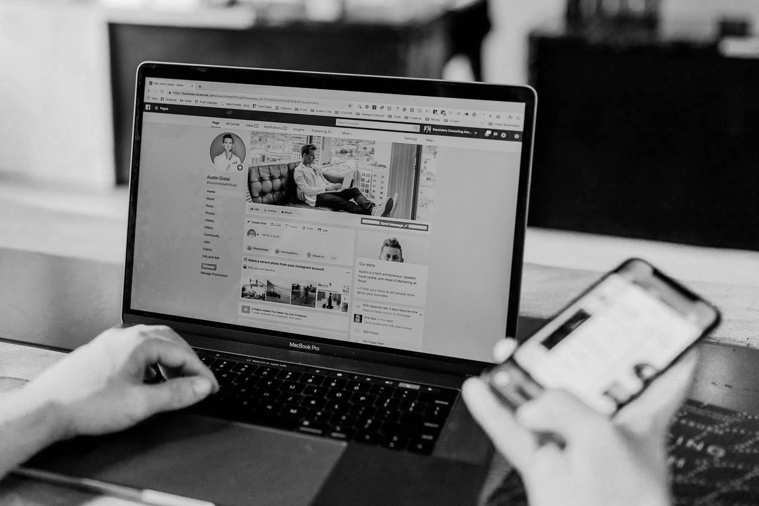 Person using a MacBook Pro displaying a Facebook profile page while holding a smartphone in a black and white setting.