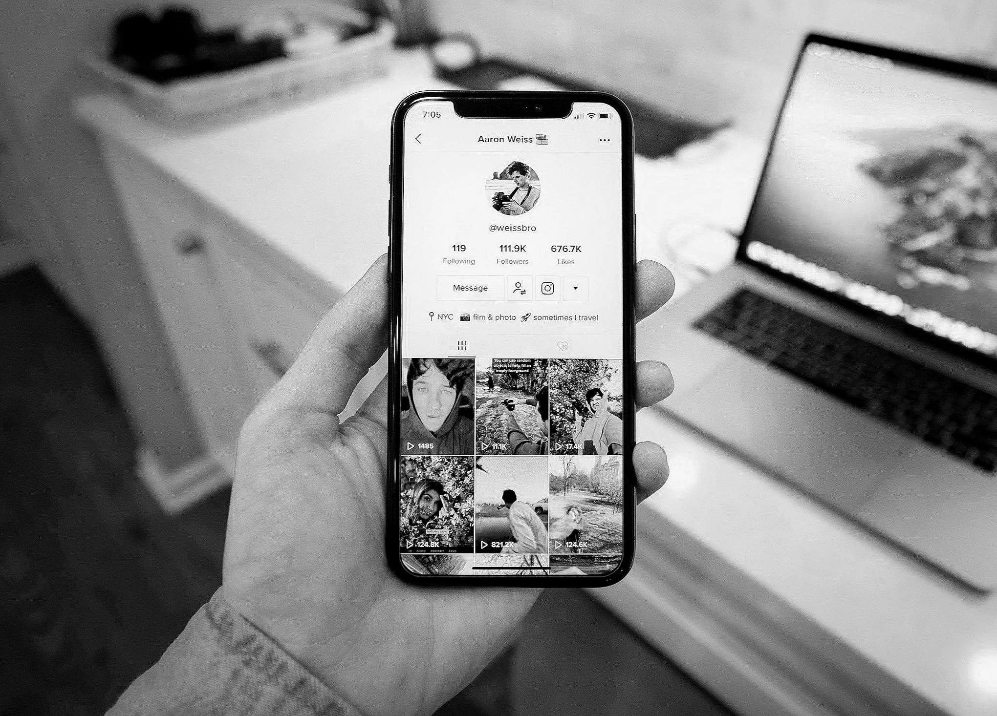 Person holding smartphone displaying a social media profile with photos and statistics; background includes a laptop and a desk.