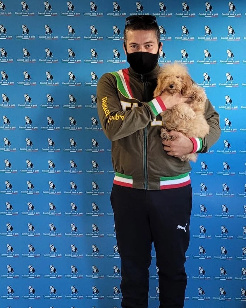 Un uomo con mascherina nera, cappellino e giacca sportiva tiene un cane peloso color beige in braccio. Lo sfondo è una parete blu con un logo ripetuto di un cane e testo.