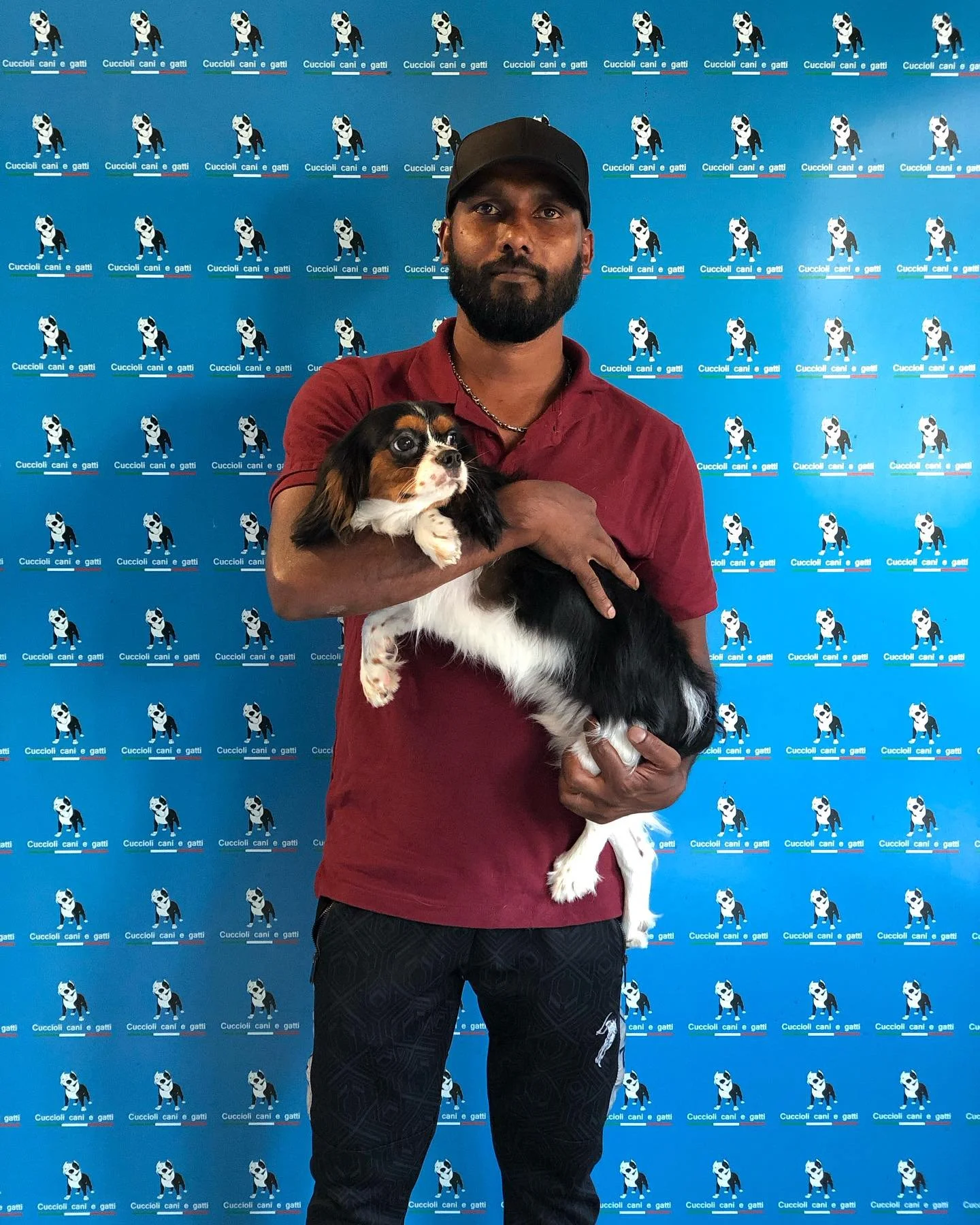 Uomo con barba nera, cappellino nero e maglietta rossa che tiene un cane di taglia piccola con pelo lungo, tricolore bianco, nero e marrone, in una sede con sfondo blu con logo di cani e gatti.