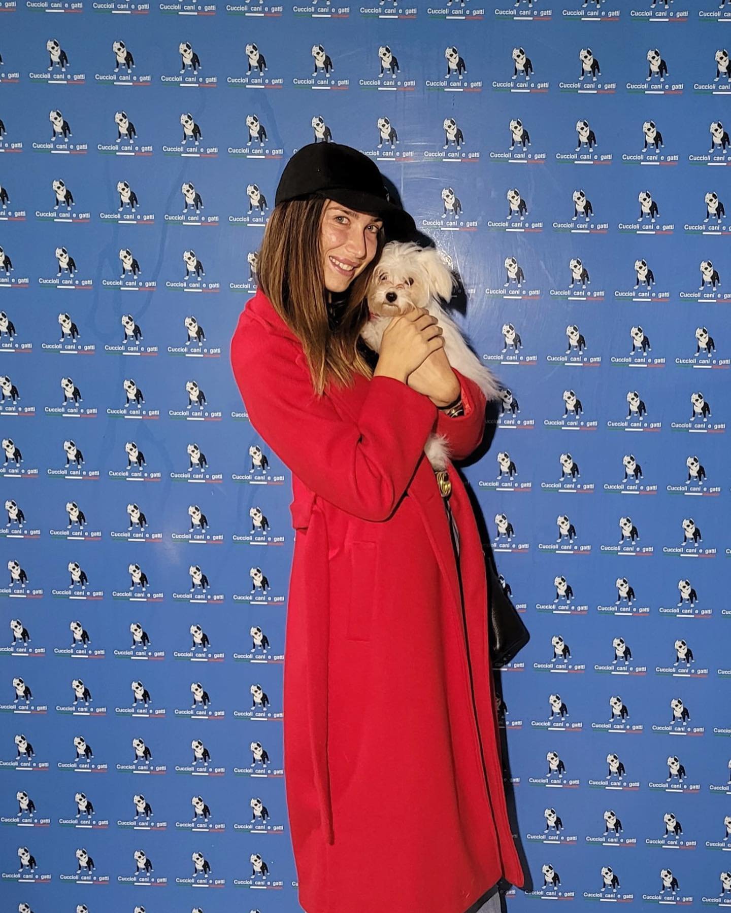 Una donna sorridente con cappello nero e giacca rossa tiene in braccio un cane bianco, davanti a uno sfondo con ripetute immagini di un cane in un cartellone pubblicitario.