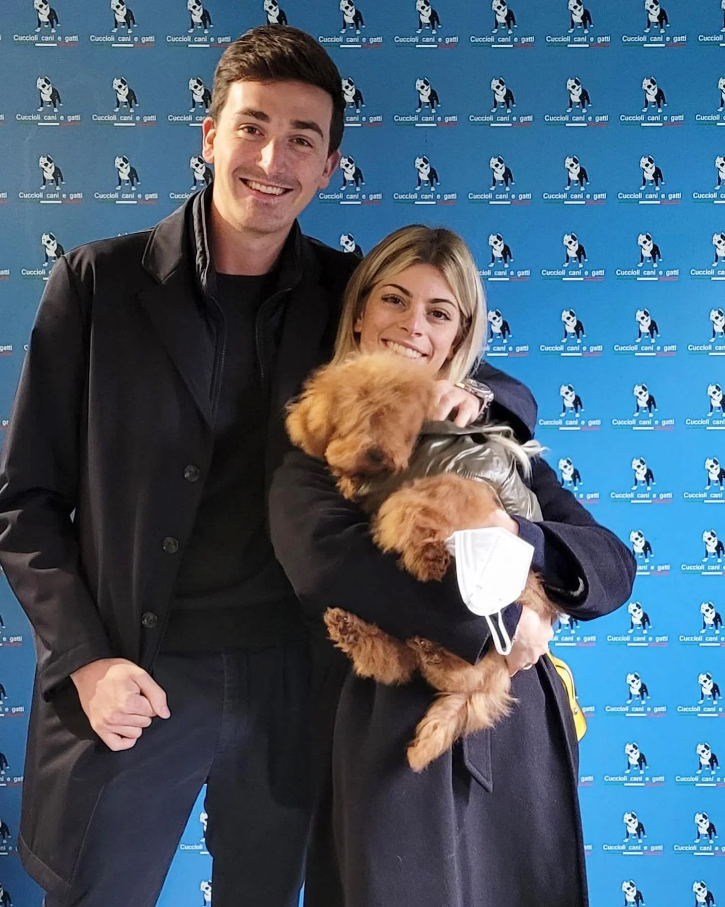 Due persone che tengono un cucciolo di cane con una mascherina, davanti a uno sfondo con il logo di un'organizzazione per cani e gatti.