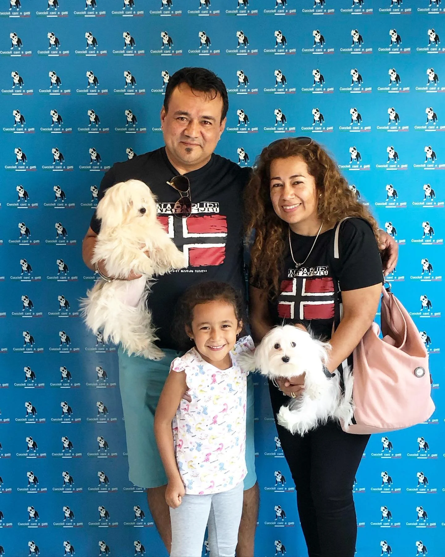 Una famiglia composta da un uomo, una donna e una bambina sorridenti, con due cani bianchi, in posa davanti a uno sfondo blu con un logo ripetuto.