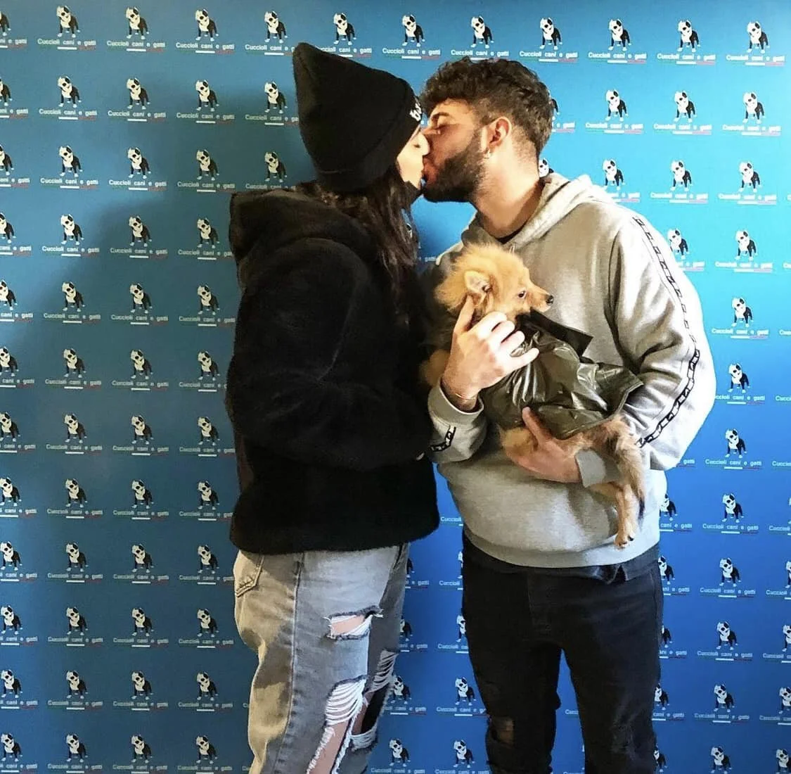 Una coppia si bacia mentre tiene un piccolo cane sulle ginocchia di uno di loro. Sullo sfondo si vede un muro con ripetute illustrazioni di un cane e il testo 'Cuccioli, cani e gatti'.