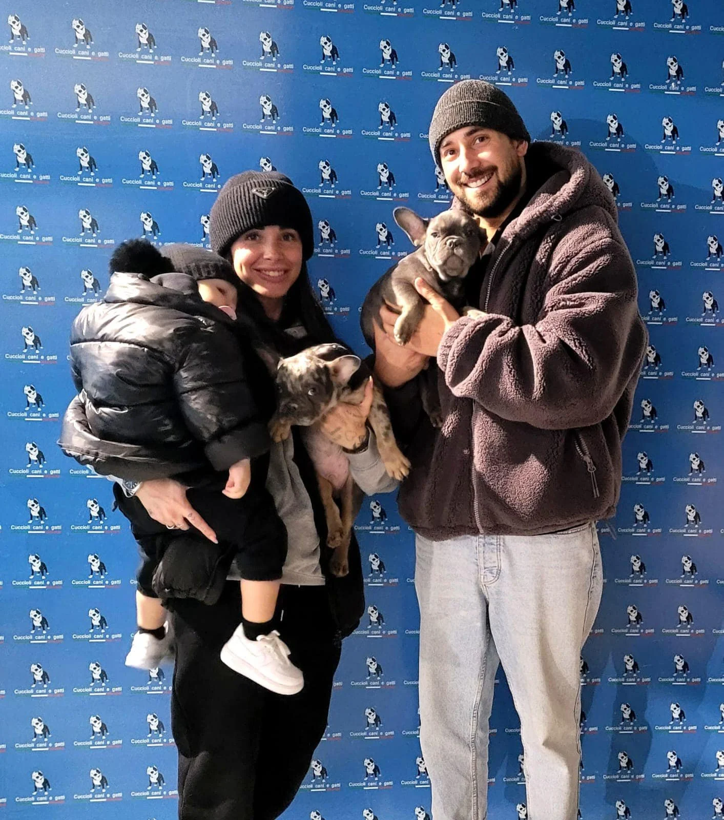 Una famiglia di quattro persone con due bambini e due cani, che sorride davanti a uno sfondo blu con disegni di cani e la scritta 'Cuccioli cani e gatti'.