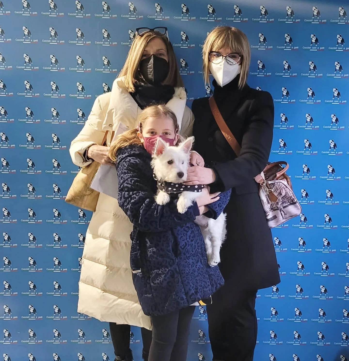 Tre donne e una bambina insieme con un cucciolo di cane bianco, tutte indossano mascherine. Sullo sfondo, una parete decorata con un motivo di cani e scritto 'Cuccioli cani e gatti'.