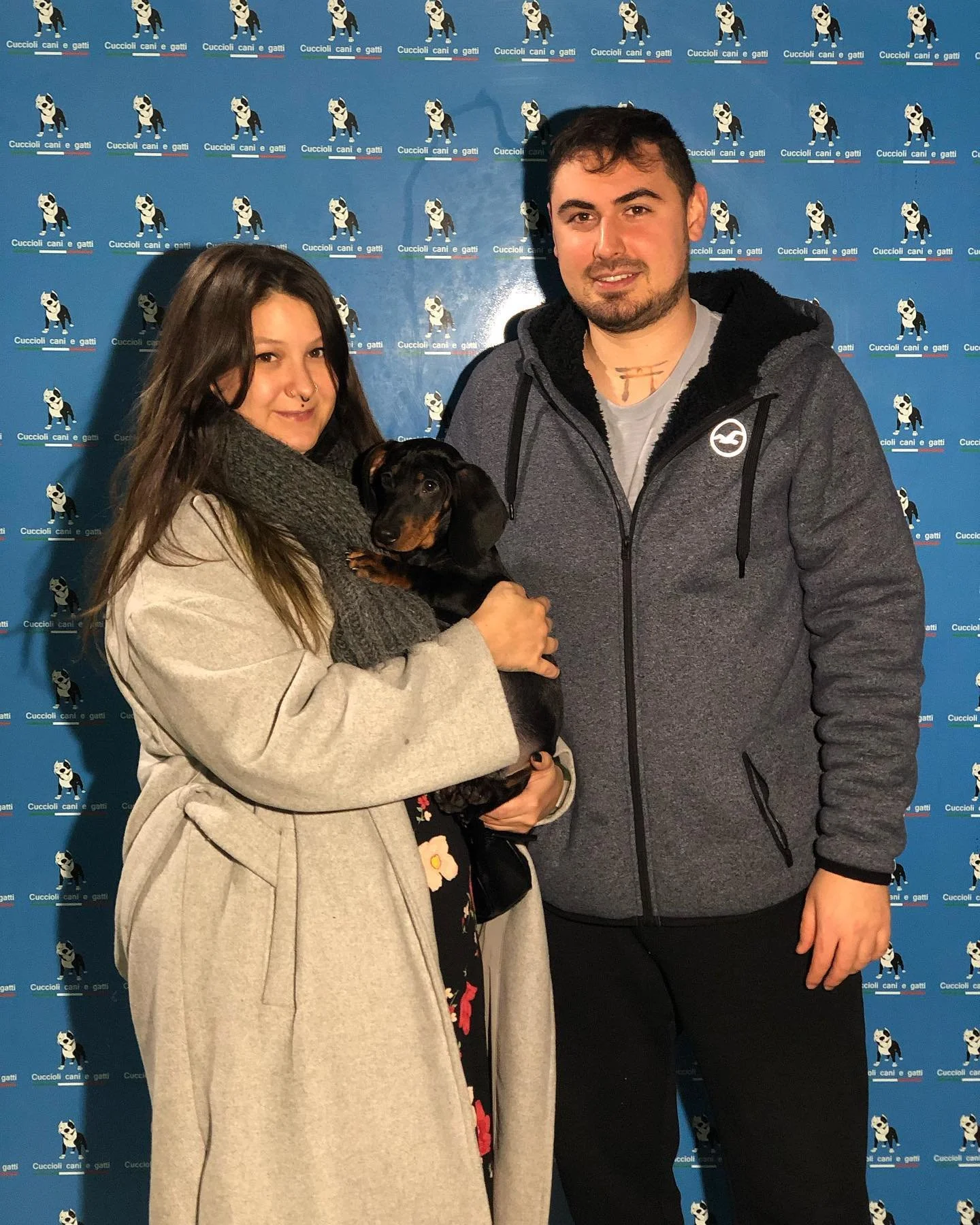 Una giovane donna con帽a beige e sciarpa grigia tiene un cucciolo di cane nero e marrone. Accanto a lei, un uomo con giacca grigia e t-shirt grigia. Sullo sfondo, un banner blu con l'immagine di un cane e scritto 'Cuccioli cani e gatti'.