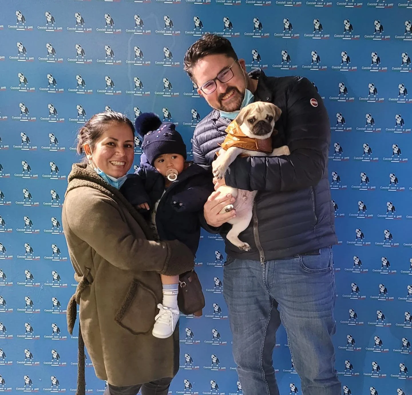 Una famiglia con un bambino e due adulti che tengono dei cani, tutti sorridenti, davanti a uno sfondo con un motivo di cani. Uno degli adulti tiene un bulldog con un giubbotto arancione.