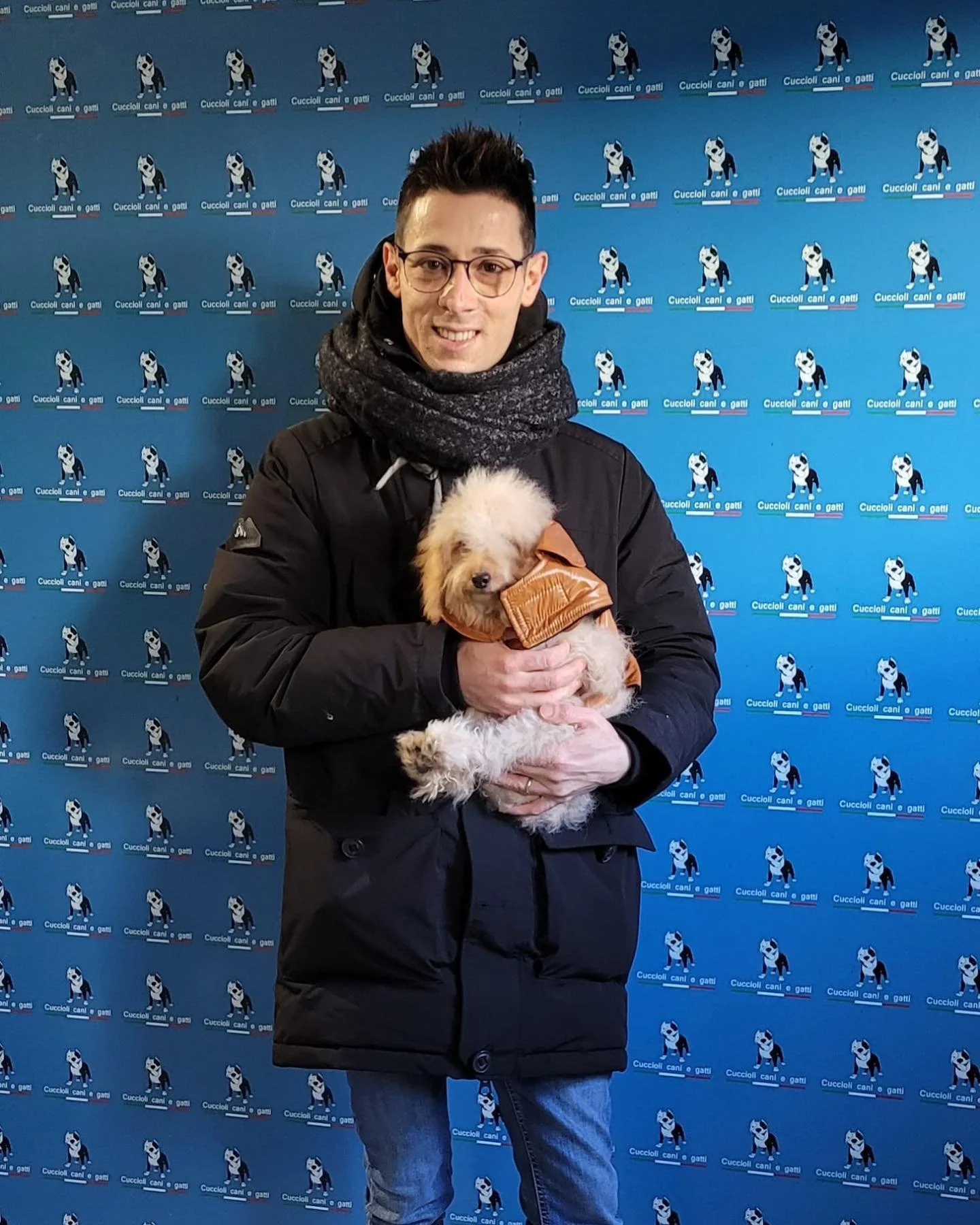 Uomo con occhiali e sciarpa che tiene un cucciolo di cane bianco e crema con un giubbotto marrone, davanti a uno sfondo con ripetute immagini di un bulldog e la scritta 'Cuccioli cani e gatti'.