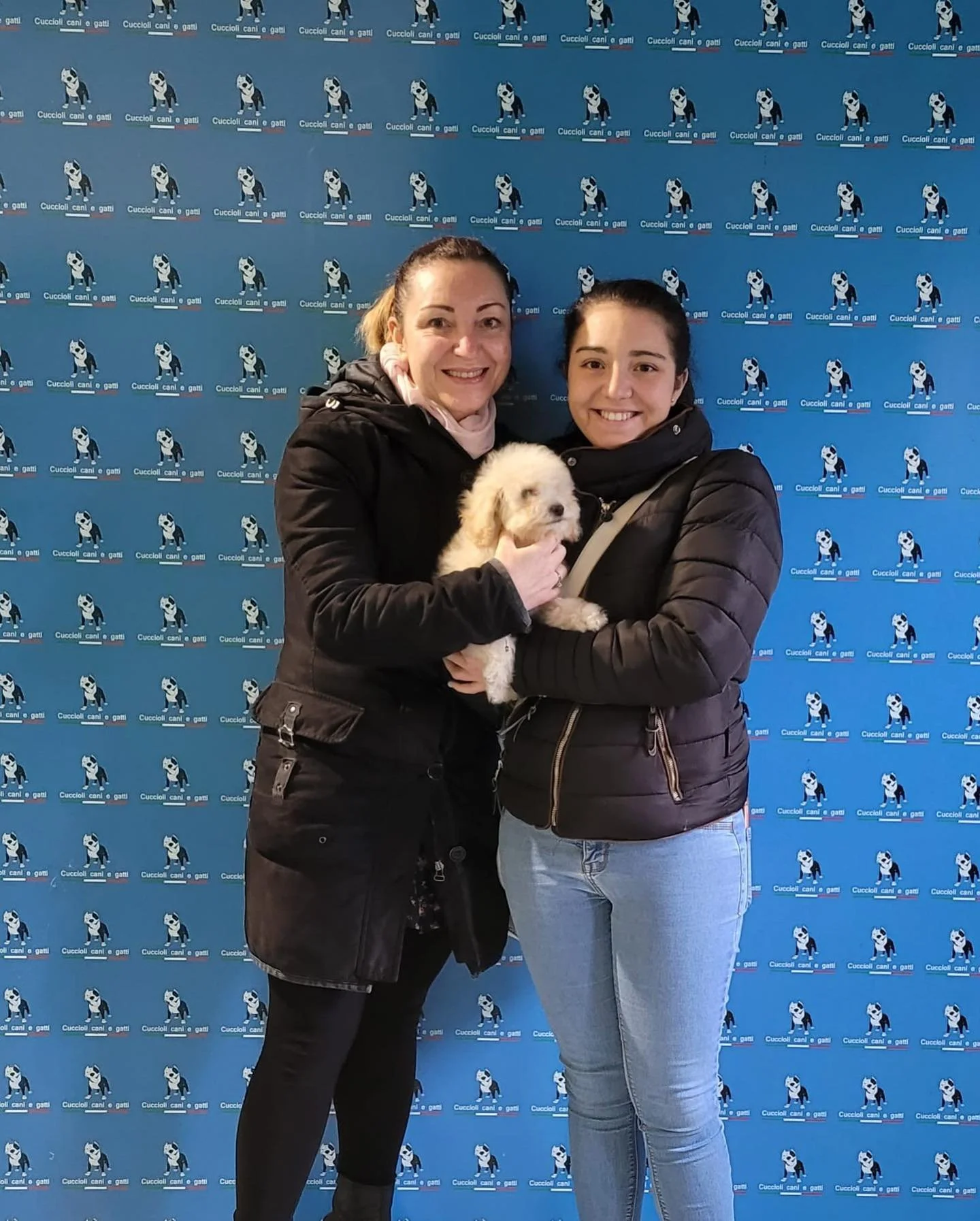 Due donne sorridenti con un cane bianco e marrone di fronte a uno sfondo blu con il logo di un canile e cani.
