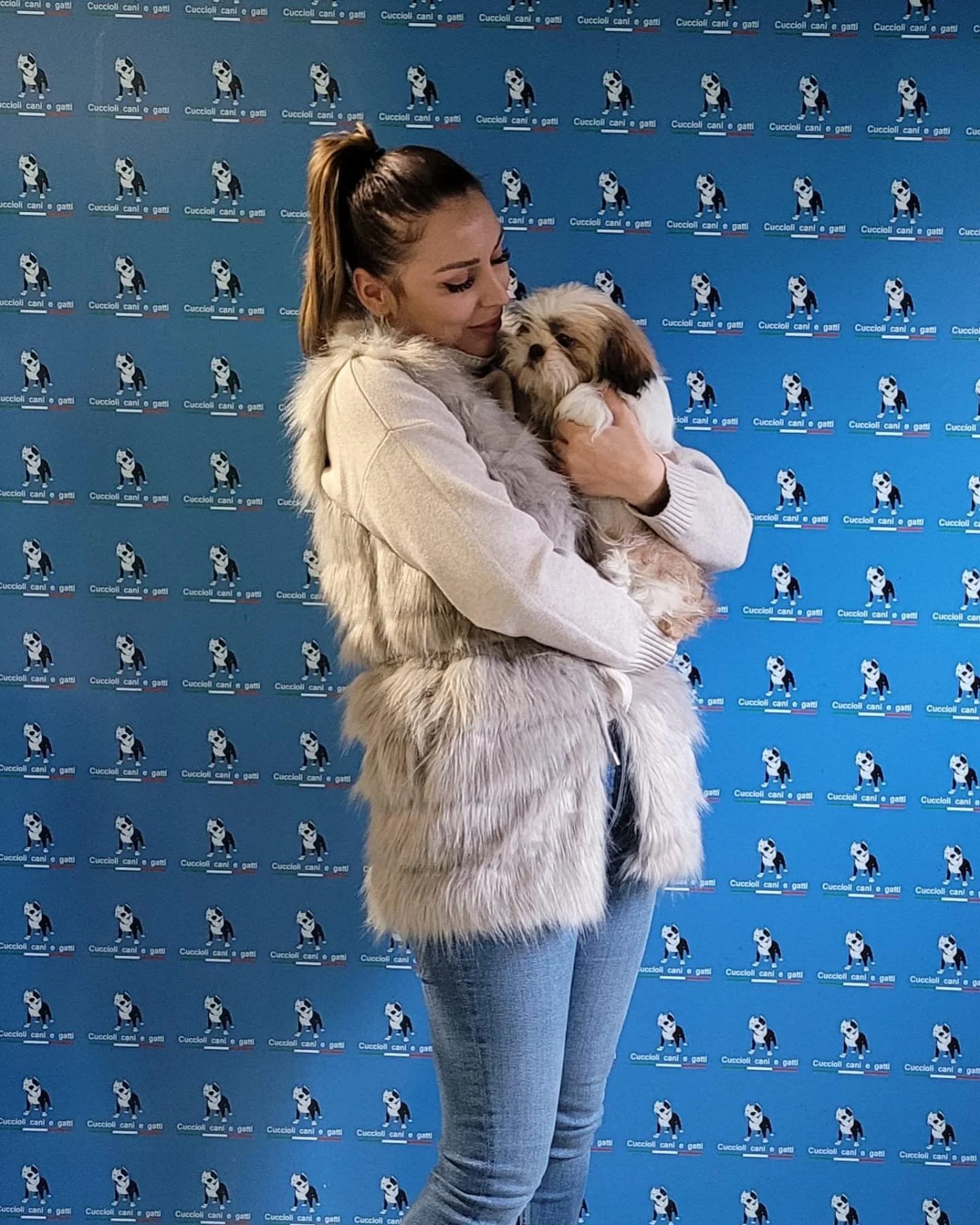 Una donna che tiene in braccio un cane di piccola taglia, sullo sfondo una parete blu con ripetuti loghi di un cane in abito e la scritta 'Cuccioli cani e gatti'.