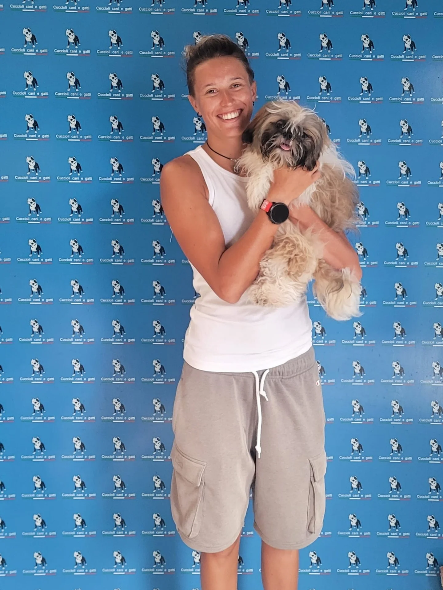 Una donna sorridente tiene un cane peloso di piccola taglia, in piedi davanti a uno sfondo con il logo di un canile.