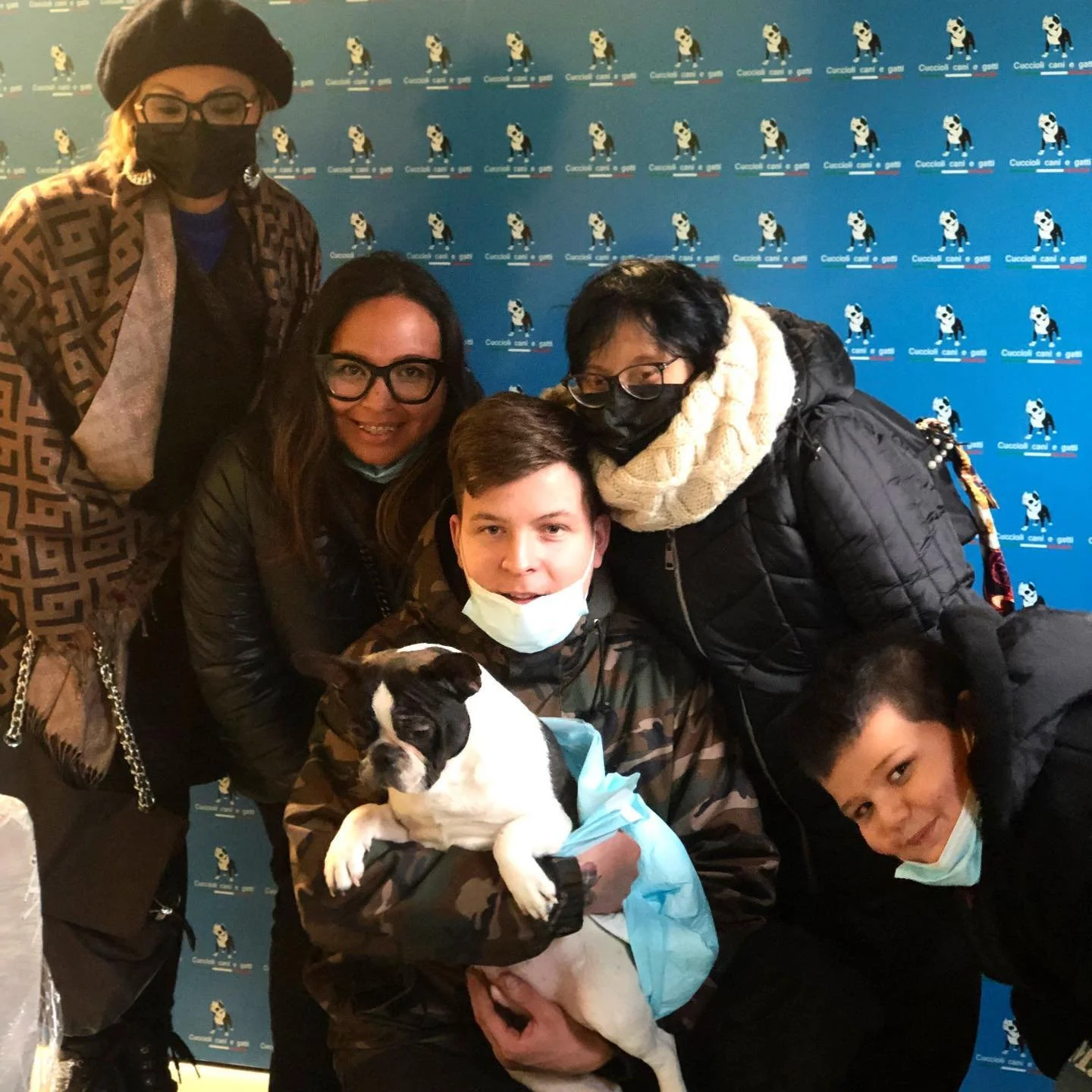 Gruppo di cinque persone con un cane dal pelo corto bianco e nero, alcune indossano maschere, in un evento con sfondo blu con logo di un cane.